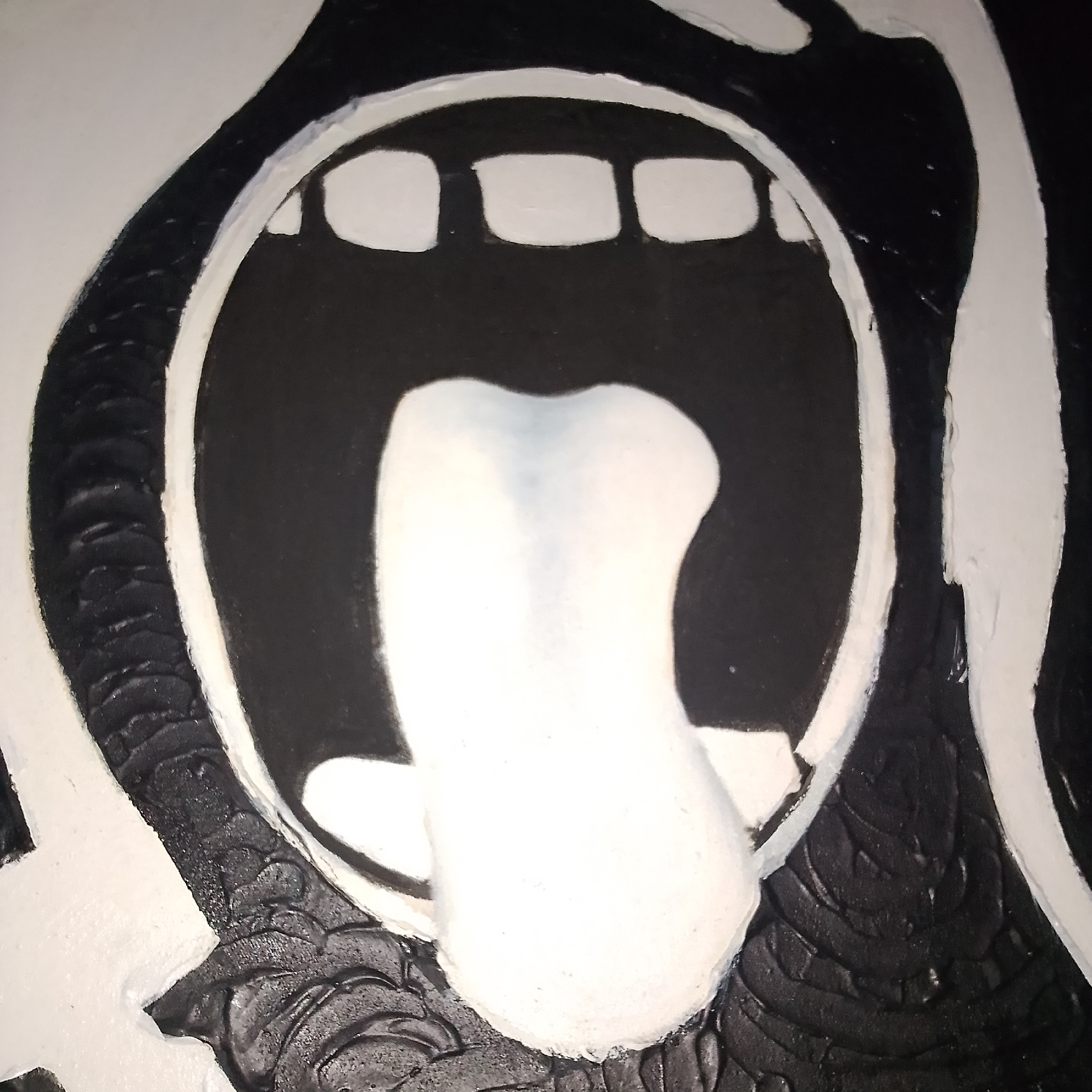 The image shows a close-up of a black and white abstract artwork, likely a mural or painting, depicting a stylized open mouth with a white tongue. The mouth is outlined in white, with black filling the interior and the background. Several rectangular white shapes representing teeth are visible within the mouth. The tongue is a large, rounded, white shape protruding from the mouth. The artwork has a textured surface, particularly in the black areas, which appear to have a rough, raised pattern. The artwork's style is bold and graphic. This artwork is located on a wall in the city of Ado, Nigeria.