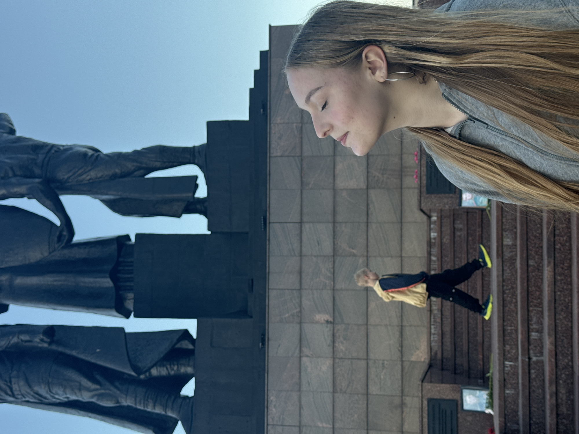 The media file displays an outdoor scene with two people and a large monument, possibly in Perm, Russia. The image is oriented vertically, with the sky occupying the top left portion and a monument taking up the rest of the frame.
In the foreground, on the left side of the image, are the legs and lower bodies of two large, dark statues, which appear to be made of bronze or a similar dark metal. The statues are depicted in a striding or standing pose, with detailed clothing folds visible.
To the right of the statues, a young woman is prominently featured, with her head and shoulders visible in profile. She has long, light brown hair and is wearing a grey hooded jacket. Her eyes are closed, and her expression suggests a moment of contemplation or peace. She is positioned in the upper right quadrant of the frame, looking downwards.
Further in the background, on the right side of the image, a smaller person, likely a child, is seen ascending a set of stone stairs. This individual is wearing a yellow jacket with blue trim and black pants, and bright neon yellow shoes. The stairs are made of reddish-brown stone with visible grain. Behind the stairs is a wall covered in large, square grey tiles.
The overall lighting suggests it is daytime, with a clear, light blue sky. The scene appears to be a public memorial or monument site, given the scale of the statues and the presence of stairs and tiled walls. No visible text is present in the image.
