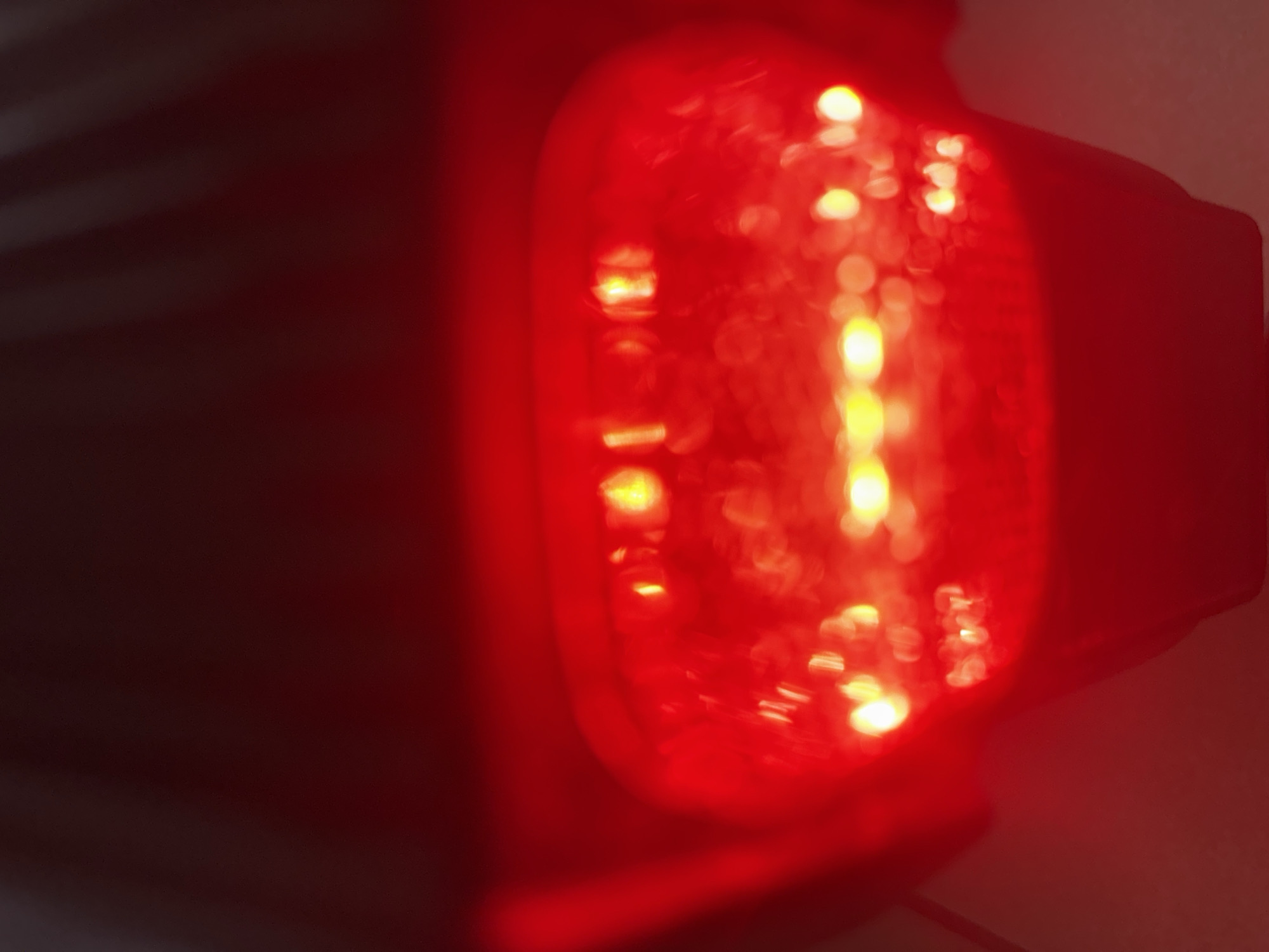 The image shows a close-up, slightly out-of-focus view of a red object, likely a light or a lens, with multiple small, bright, circular illuminated elements visible within it. The overall color palette is dominated by deep reds and dark shadows, with some brighter, yellowish highlights emanating from the illuminated points. The background is dark and indistinct, with some subtle linear patterns visible in the shadowed areas on the left, which could suggest a textured surface or a light source casting shadows. The texture of the red material appears somewhat smooth and possibly translucent, allowing the internal lights to glow through. There is no discernible activity or event taking place, as the image is a static close-up of an object. No text is visible, and there are no specific cues about the time of day, weather, or location, other than the provided context of Anduze, France, which cannot be visually determined from the image itself. The lighting suggests the object itself is illuminated, possibly from within, creating a warm and somewhat abstract visual effect.