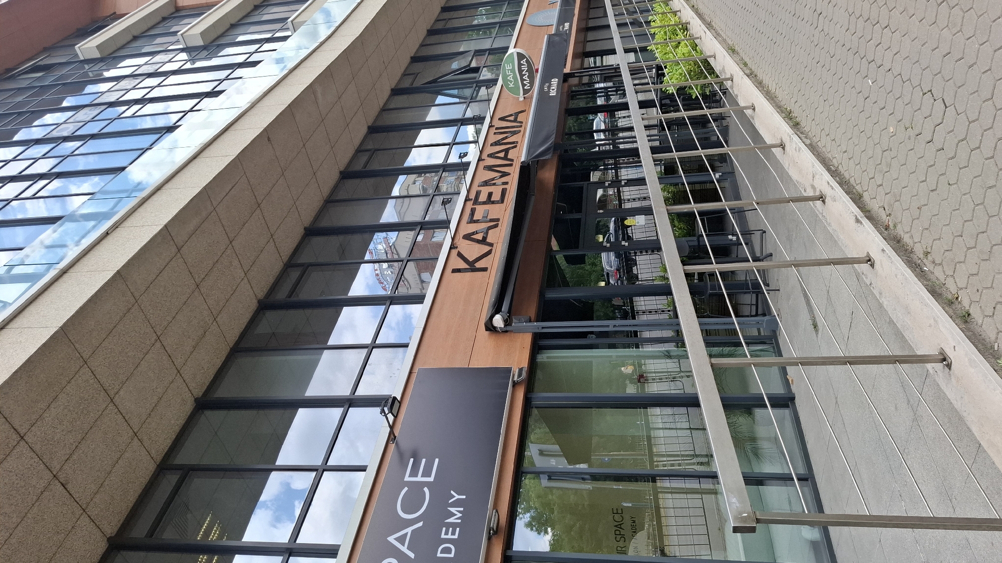 A street-level exterior shot shows a modern building with large glass windows, reflecting a cloudy sky. A prominent sign on the building reads "KAFÉMANIA" in black lettering. Below this, another sign says "KAFFÉ MANIA" in a green oval, and beneath that, a black awning displays "CAFE RICHARD". A dark sign to the left reads "SPACE ACADEMY". The building is located on a sidewalk with a metal railing. The scene is set in Sofia, Bulgaria.