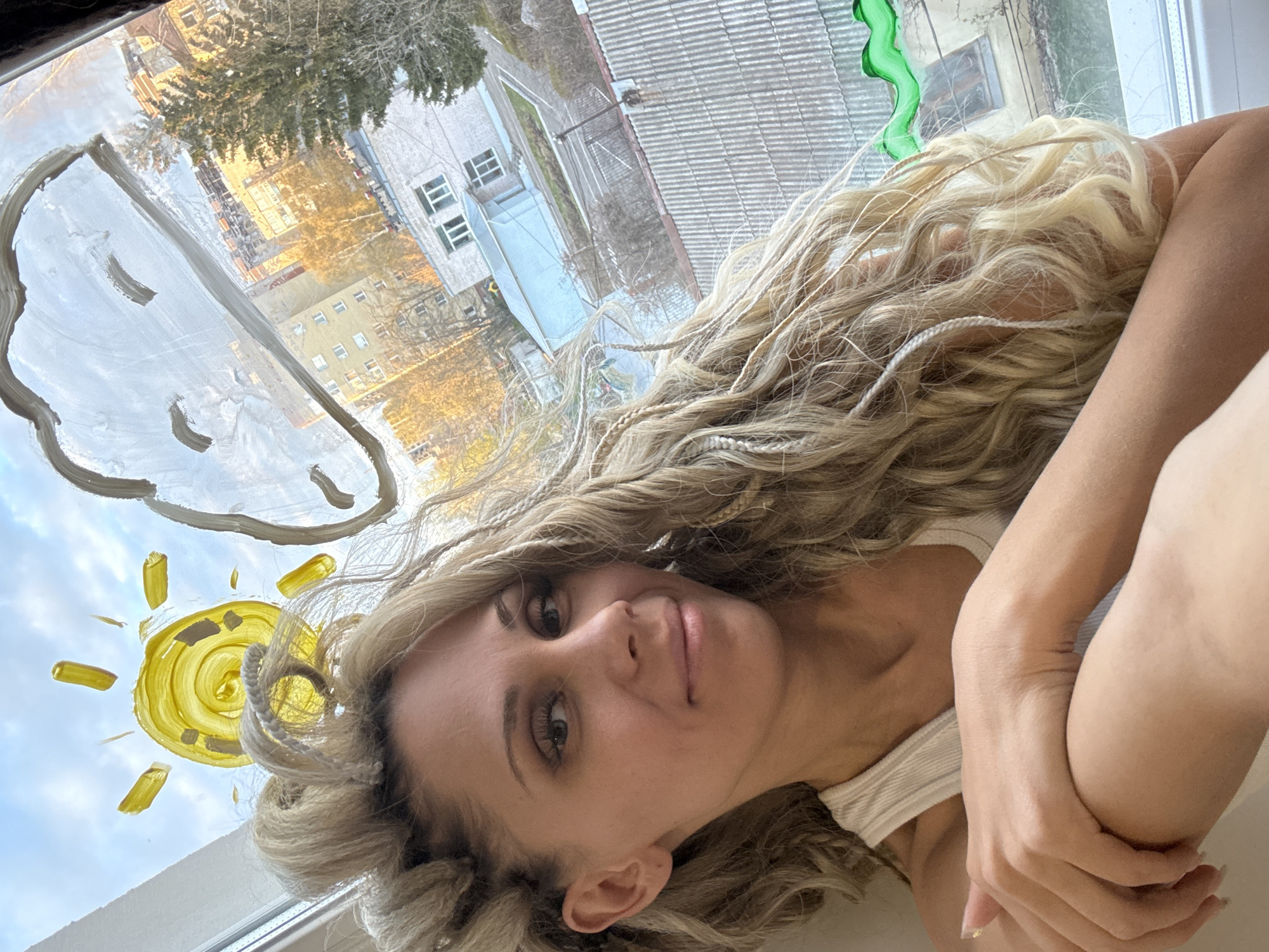 The media file features a young woman posing indoors next to a window, looking directly at the camera. She has long, wavy blonde hair with several thin braids or dreadlocks interwoven, some of which are lighter in color. Her expression is calm, with a slight hint of a smile. She wears a light-colored, possibly white, ribbed top and has her arms crossed in front of her.
The window glass itself is adorned with drawings: a large, light-colored cloud shape with simple eye-like features and a mouth, and a bright yellow sun with radiating lines, positioned lower and to the left. A small, wavy green object, possibly a decoration, hangs near the top right edge of the window.
Through the window, an urban landscape is visible. It features several residential buildings, including a multi-story yellow building illuminated by warm, golden light, suggesting either early morning or late afternoon. Another grey building with visible windows is also present. Leafless trees or trees with sparse foliage can be seen, hinting at a colder season like late autumn, winter, or early spring. A corrugated grey roof of another structure is also visible. The sky beyond the drawings appears mostly clear and blue.
The woman appears to be taking a selfie or being photographed, likely enjoying the light or the playful drawings on the window. The scene suggests a relaxed moment in a residential setting in Rusanovo, Russia. There is no visible text in the image.