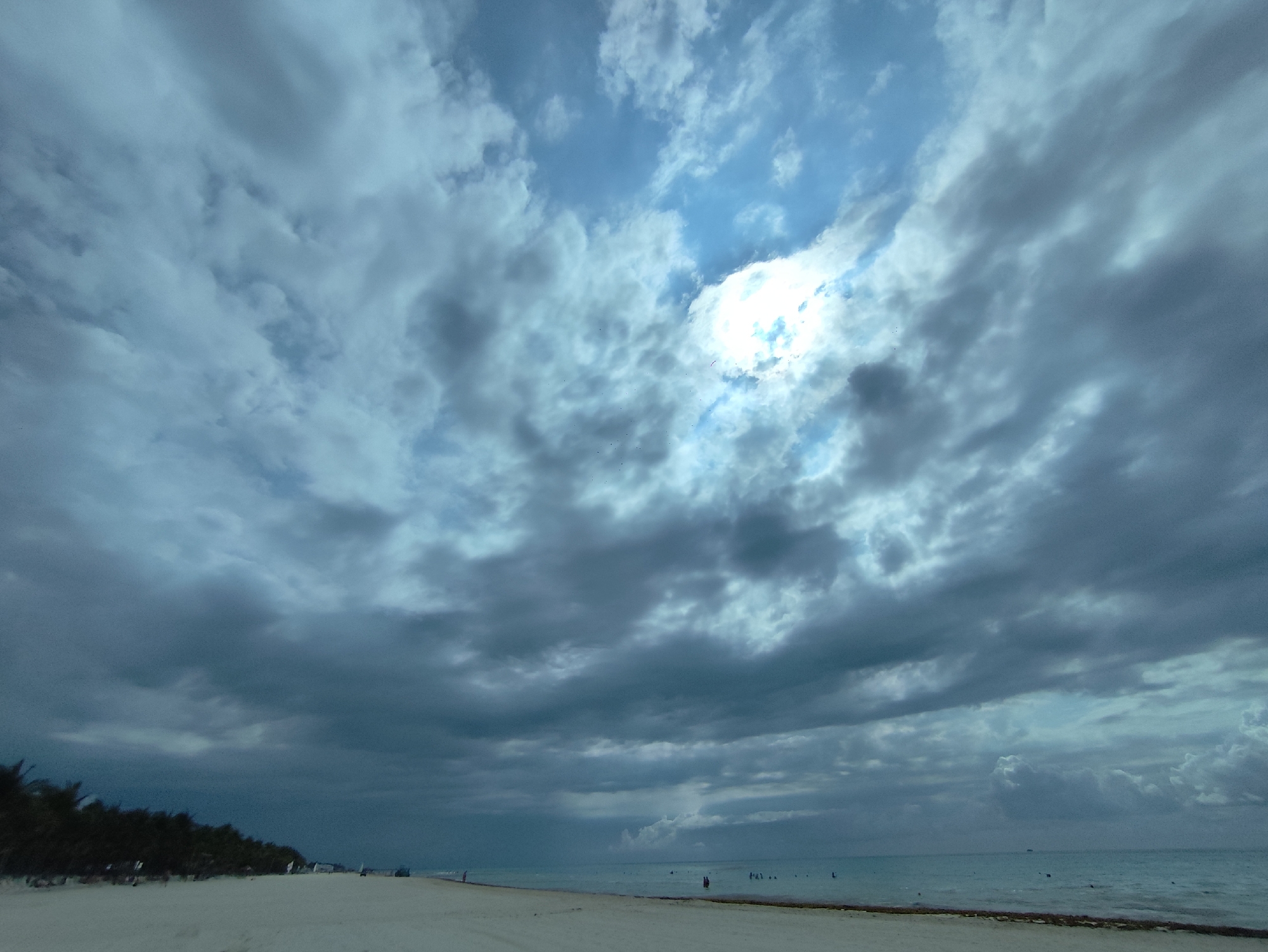 The image shows a beach scene at Playa del Carmen, Mexico, under a cloudy sky with patches of blue. The foreground features a wide expanse of white sand beach. Along the left edge are some low trees and vegetation. In the distance, the ocean meets the sky at the horizon. Several people are visible in the water, and a few are scattered along the beach. The ocean has a greenish-blue hue. There is seaweed on the sand where the beach and ocean meet. The clouds dominate the sky, ranging from light gray to dark gray, with a bright spot where the sun is trying to break through.