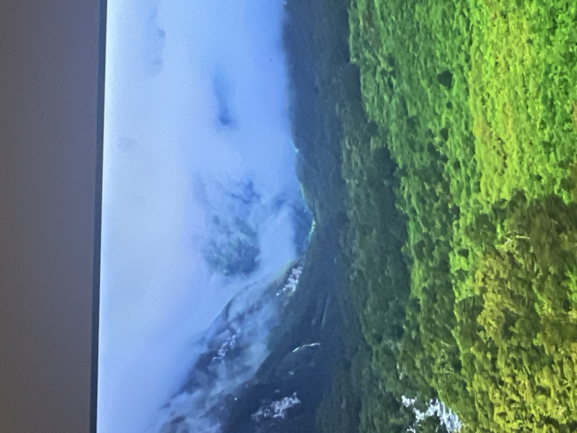 The image shows a breathtaking landscape of a lush green valley nestled between mist-shrouded mountains. A thin waterfall cascades down the side of one of the mountains, disappearing into the dense foliage below. The sky is a soft blue, with wisps of white clouds clinging to the mountain peaks, creating a serene and majestic atmosphere. The vibrant green of the trees and the muted blues and grays of the mountains and mist create a striking contrast. This image does not depict any location in the city of Casablanca, Morocco.