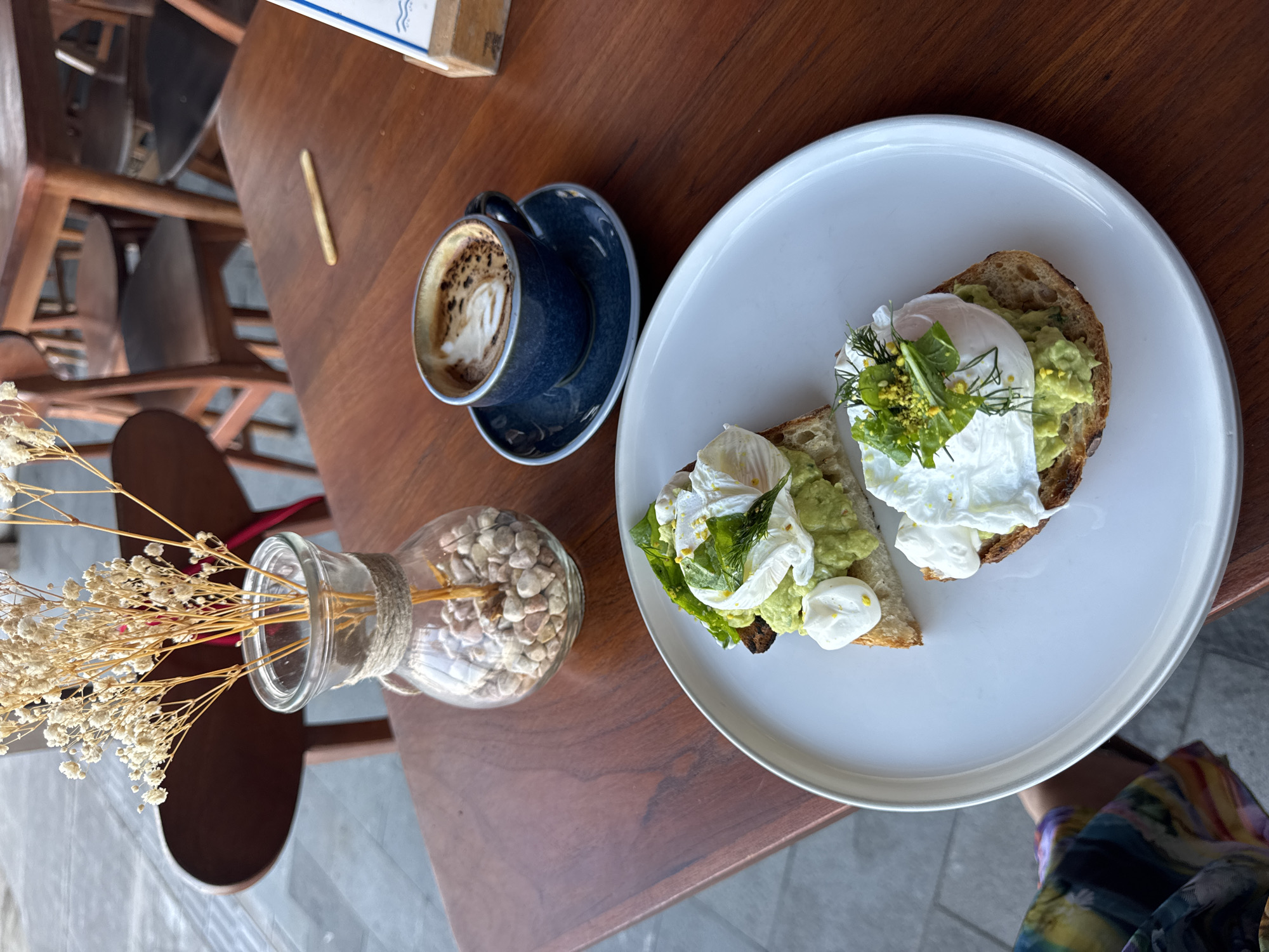 The image captures a top-down view of a meal set on a polished wooden table, likely in an outdoor or semi-outdoor cafe in Kuta, Indonesia.
The primary subjects are food and beverages: a white oval plate holds two pieces of avocado toast. Each toast piece is generously topped with mashed avocado, a perfectly poached egg, fresh green herbs like dill and what appears to be spinach or lettuce, and fine yellow sprinkles, possibly spice or pollen. To the left of the plate, a dark blue ceramic cup with a matching saucer contains a coffee drink, possibly a latte or cappuccino, adorned with visible latte art.
Next to the coffee, a clear glass vase, wrapped with a rustic twine accent, holds dried white flowers resembling baby's breath and thin, dry straw-like stems. The vase's base is filled with small, light-colored pebbles or stones. A thin, light-brown stick, possibly a biscuit or breadstick, lies on the table between the coffee and the blurred background.
In the background, out-of-focus wooden chairs suggest a seating area. A small portion of a wooden box or crate is visible in the top left corner, displaying an unreadable blue logo or text that appears to include stylized waves. The bright, natural lighting suggests it is daytime. Part of a person's colorful, patterned clothing is visible in the bottom right corner, indicating someone is seated at the table, about to enjoy or having just enjoyed the meal. The overall scene conveys a relaxed and pleasant dining experience.
