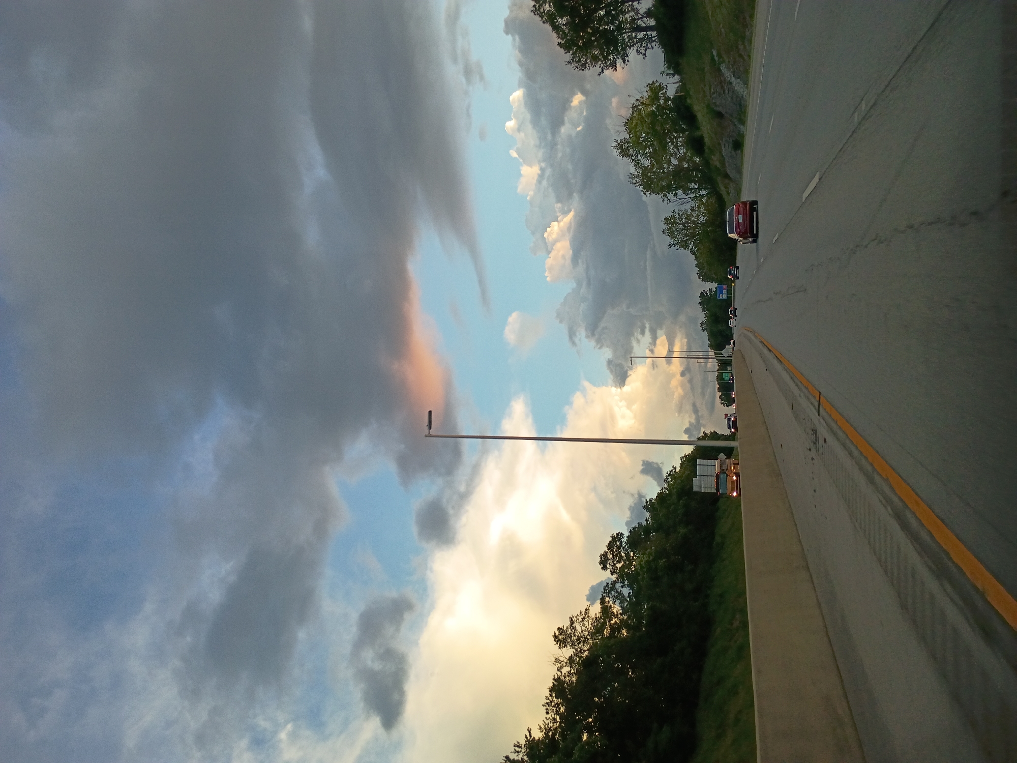 The image depicts a view from a vehicle traveling on an elevated highway, possibly Interstate 75 North near Lexington, Kentucky, USA. The sky is dramatic, filled with a mix of dark storm clouds and brighter, cumulonimbus clouds tinged with sunlight. A tall lamppost stands prominently on the right side of the highway. The highway itself has multiple lanes, with traffic moving in the same direction. Several cars are visible, including a red sedan in the foreground and a yellow truck further ahead. Trees and dense foliage line the embankment to the left of the highway, while to the right, a grassy incline with scattered trees rises. The sun is low in the sky, casting a warm glow on parts of the clouds and illuminating the scene.