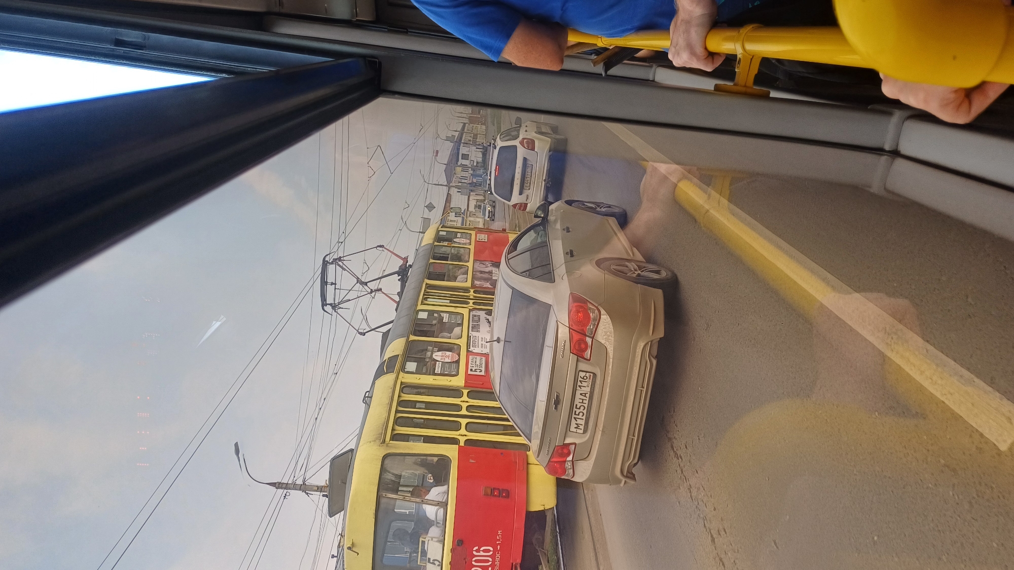 The image shows a view from a bus window in Izhevsk, Russia. A yellow and red tram is visible on the left, with the number 206 on its side. Behind the tram, a beige sedan with a Russian license plate M155HA 116 is driving on the road. Further down the road, a white SUV is also visible. The sky is overcast. The interior of the bus is visible on the right side of the image, with yellow handrails and a person in a blue shirt holding onto one of them. The overall scene depicts a typical street view in a Russian city with public transportation and road traffic.
