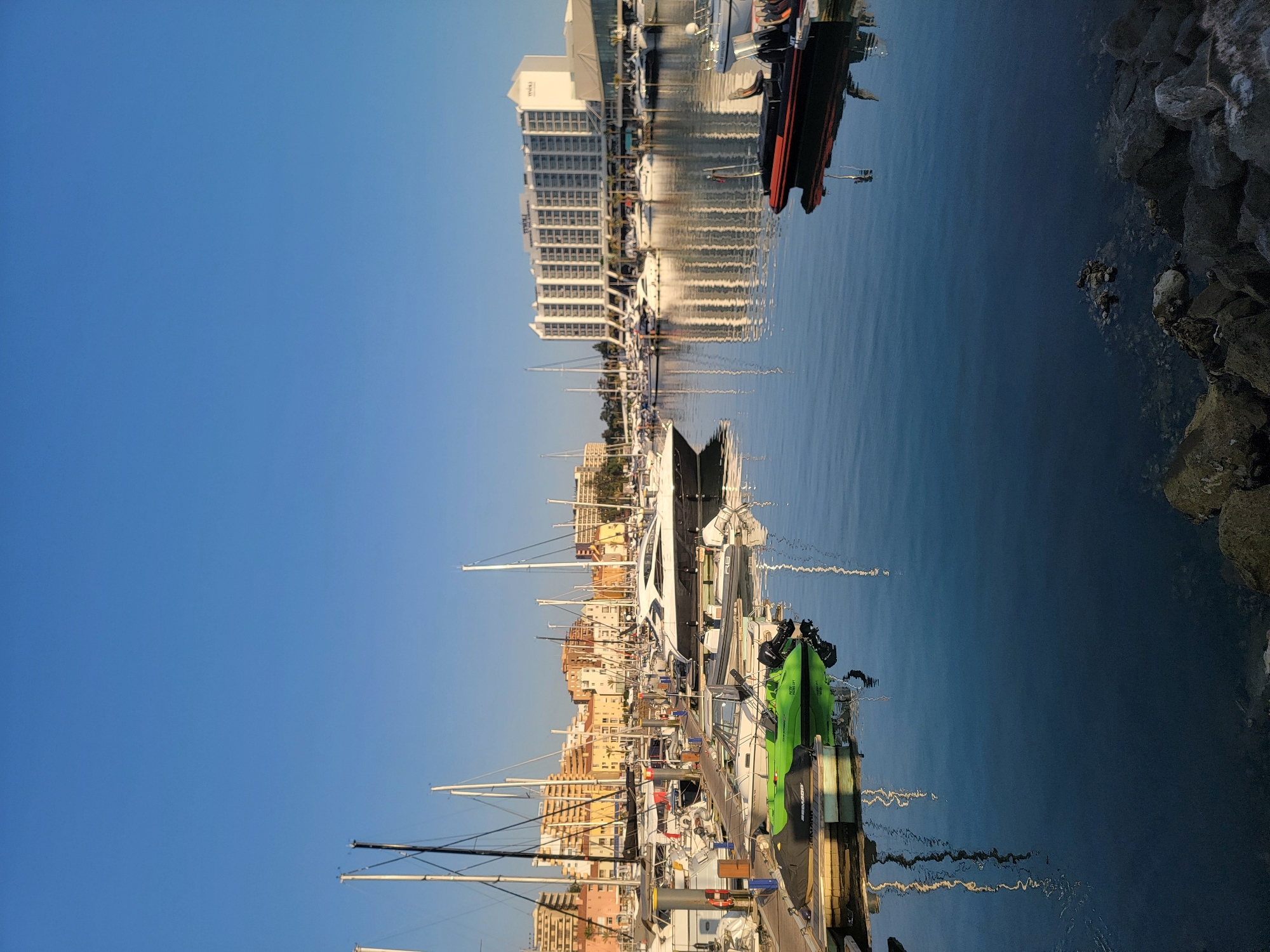 The image depicts a marina in Quarteira, Portugal, under a clear blue sky. Numerous boats are docked, ranging from sleek yachts with white hulls to smaller speedboats and a bright green jet ski. The boats are arranged in rows along wooden jetties. In the background, a large white hotel with many windows stands prominently, reflecting on the calm water of the marina. Several other buildings, appearing to be apartments or other hotels, are visible further down the coastline, with terracotta roofs and balconies. The foreground shows the calm, reflective surface of the water with some rocks visible at the bottom right corner. There are no people visible in the image. The overall scene is one of a peaceful coastal resort town.