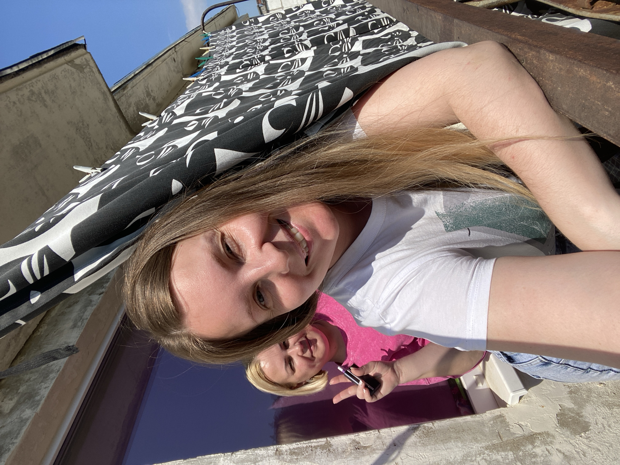 A young Caucasian woman with blonde hair is smiling at the camera, taking a selfie. She wears a white t-shirt and blue jeans. Reflected in a dark, presumably reflective surface below her is a second woman, also Caucasian, with blonde hair, wearing a pink shirt and holding a small black rectangular object. She is making a peace sign with her fingers. A black and white patterned sheet or fabric hangs overhead, clipped to a line with clothespins. In the background, architectural details suggest an urban environment, potentially in Noviy, Russia, with parts of buildings visible. A rusty metal structure is positioned to the right of the first woman. The overall lighting suggests an outdoor, sunny day.