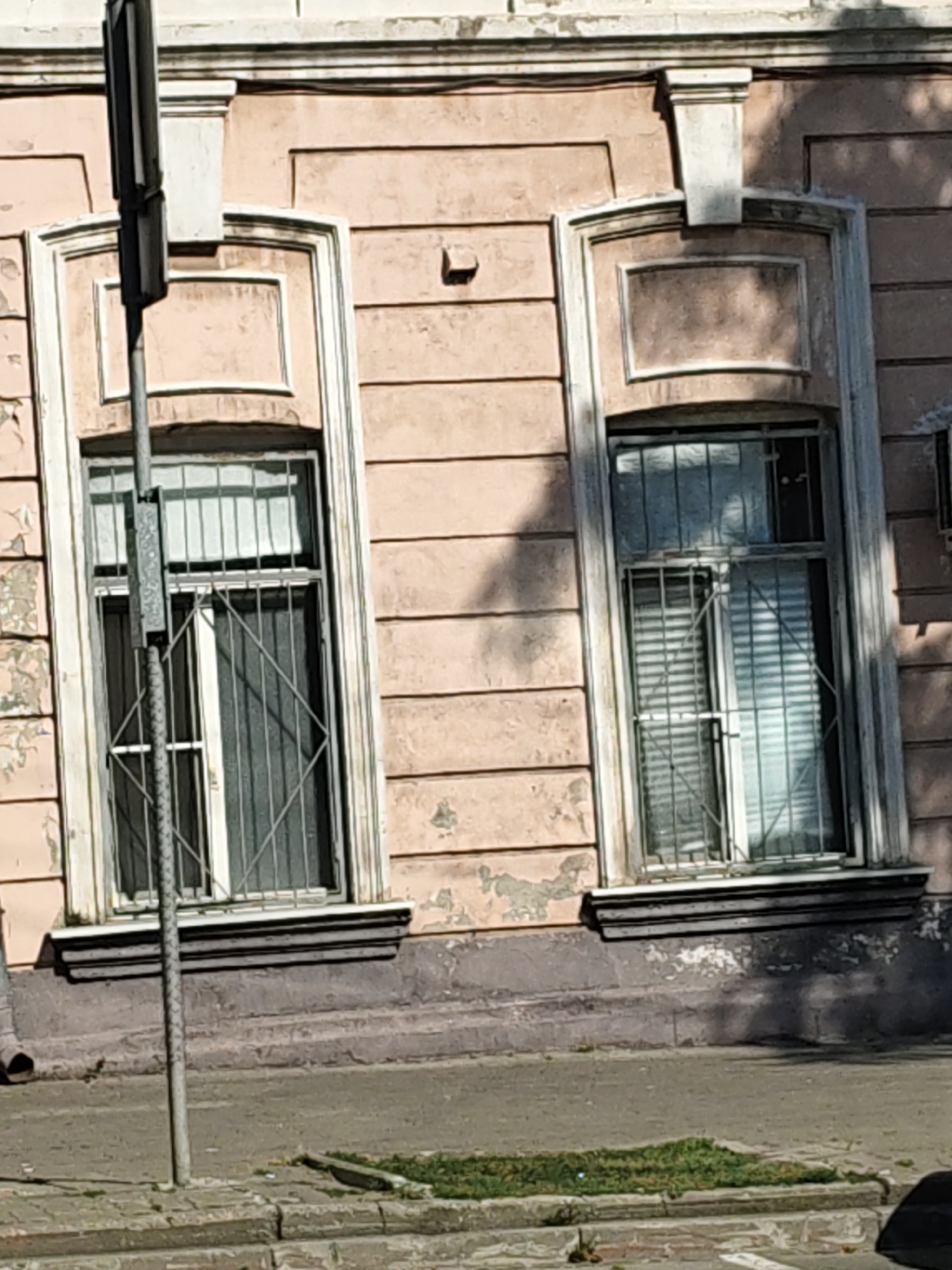 The image shows the facade of an old building with two tall, narrow windows. The building's exterior wall is painted a faded pink and appears to be made of stucco with horizontal texturing. Both windows are protected by metal bars and have white sills. The window on the left is partially obscured by a metal pole, likely a street light or signpost, which is positioned in front of it. The window on the right is mostly visible, revealing blinds or shutters behind the bars. Below the windows, there is a dark-colored base or plinth with peeling paint. The ground in front of the building consists of a sidewalk and a small patch of grass. A drainpipe is visible on the left side of the building. The overall scene suggests an urban environment in a state of some disrepair, typical of older architecture. The building's architectural style, particularly the decorative elements above the windows and the horizontal texturing of the facade, is consistent with buildings found in Rostov-on-Don, Russia, a city known for its historical architecture.