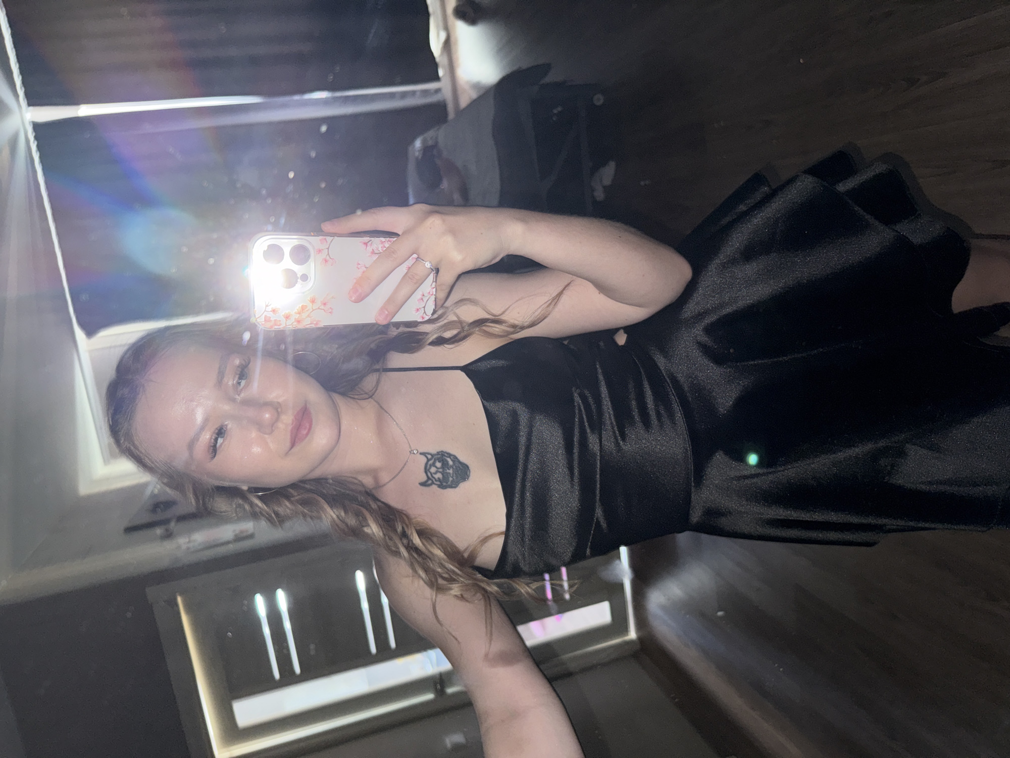 A young woman with long, wavy blonde hair is taking a selfie in a mirror. She is wearing a black satin slip dress and a necklace with a pendant. A ring is visible on her right hand, which is holding a smartphone with a floral case. The phone's flash is on, creating lens flare in the image.
The background appears to be an indoor setting, possibly a room with unfinished walls and a wooden floor. There are reflections in the mirror, including what looks like furniture and a window with vertical blinds. The lighting suggests it might be evening or a dimly lit room. The woman has a relaxed expression. No visible text is present in the image.