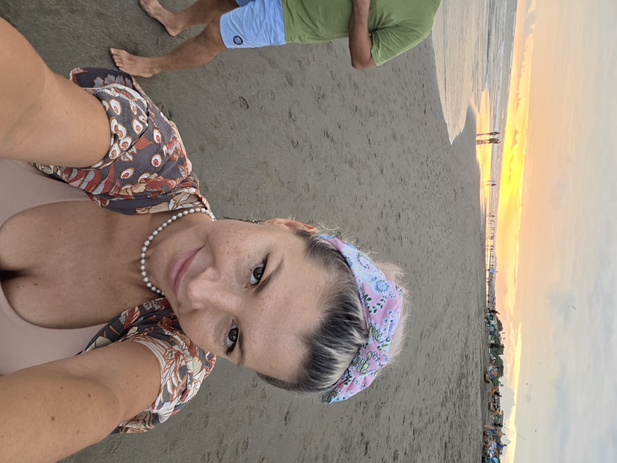 This is a selfie taken on a beach in North Kuta, Indonesia, during sunset. The foreground is dominated by the woman taking the photo, who is smiling and looking at the camera. She has blonde hair tied back with a pink and white floral bandana, and she is wearing a pearl necklace and a patterned cover-up over a pink bikini top. To the left of the frame, the legs of a person wearing a green t-shirt and blue shorts are visible. The background shows a sandy beach with waves rolling in, and the sky is painted with warm sunset colors of orange and yellow. Several people are scattered along the beach in the distance, and some are near beach umbrellas and structures.
