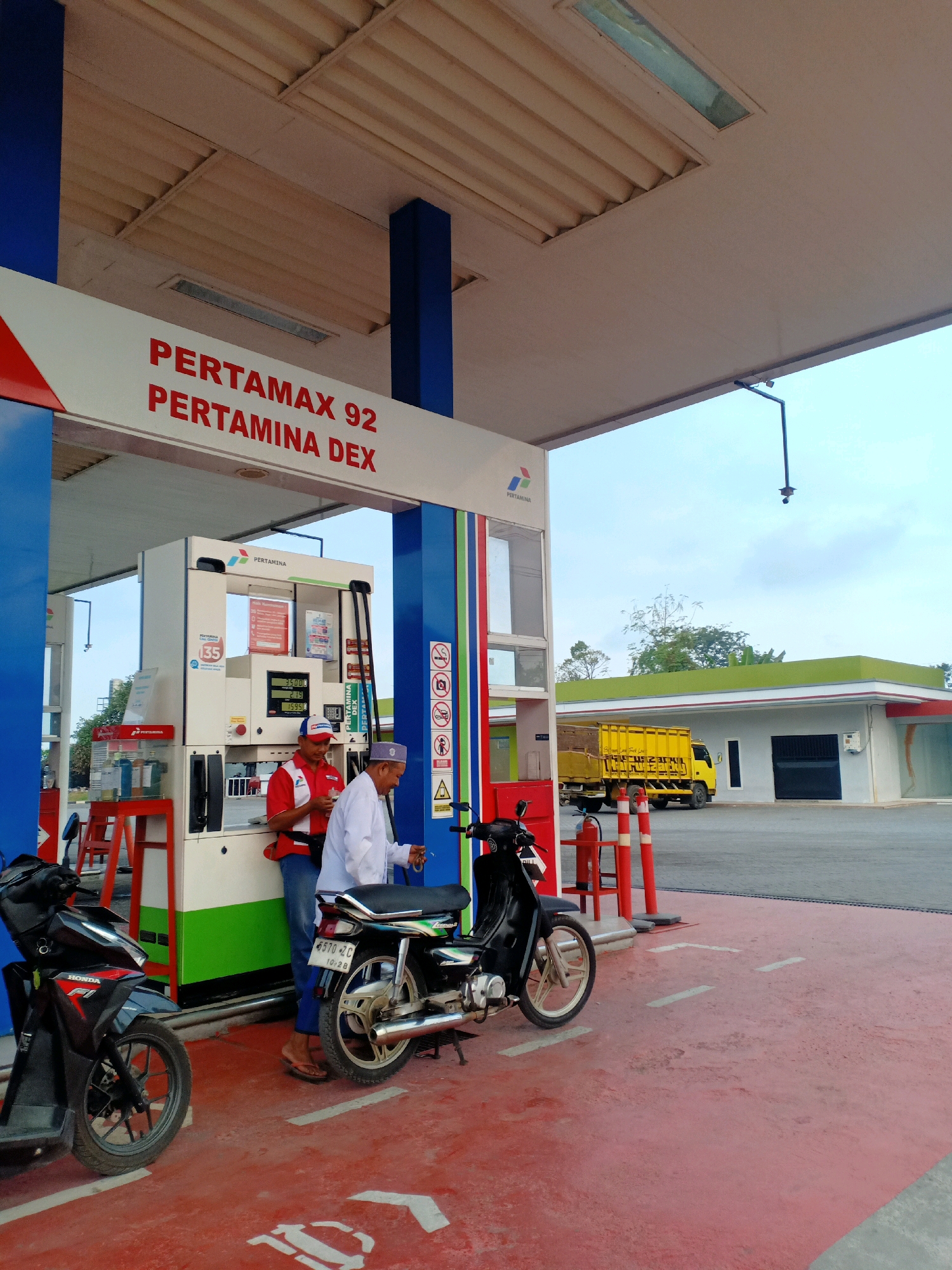 The image depicts a gas station with a red ground. In the foreground, two motorcycles are parked. To the left, a black scooter with red accents is visible. To the right, a black motorcycle with silver accents and a license plate reading "5570 JC 10:26" is parked. Two men are standing near a Pertamina gas pump. One man, wearing a red shirt with a white collar and blue jeans, appears to be a gas station attendant and is holding a cigarette. The other man, dressed in a white robe and cap, is facing away from the camera. The gas pump displays prices for "PERTAMAX 92" and "PERTAMINA DEX." In the background, a yellow truck with a cargo bed is parked near a building with a green roof. The sky is clear and bright. The overall scene suggests a typical day at a roadside gas station.
