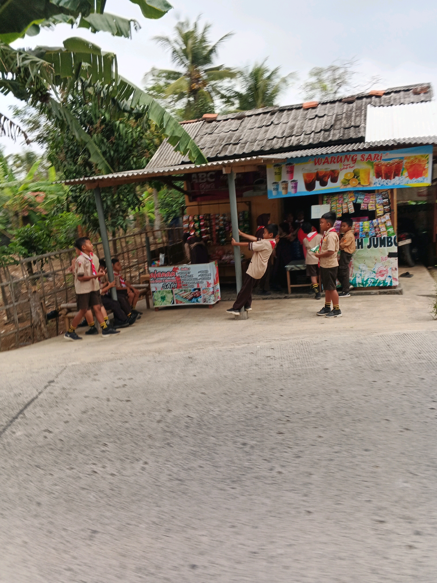 The image shows a street scene in Jangrana, Indonesia, with a small roadside shop and several young people in scout uniforms. The shop, possibly a warung, has a corrugated metal roof and a banner advertising drinks. Shelves display various packaged goods. On the left, a wooden fence separates the road from vegetation and trees. Several boys in scout uniforms are present. One boy is standing near a pole, leaning back and holding it with his hands, with his left foot on a raised object. Other boys are sitting or standing near the shop. The road surface is concrete with some cracks. The overall atmosphere appears casual and outdoor.