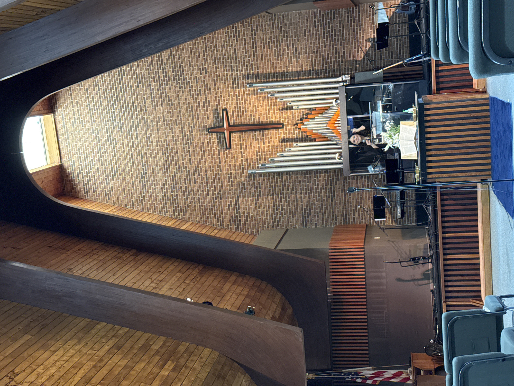 A view from the interior of a church, with an eye-level view of the wooden slatted floor, a curved wooden beam going towards the ceiling, and a brick wall with a cross and organ pipes above the stage. The pastor is at the podium.
The church appears to be located in Columbus, Ohio, USA.