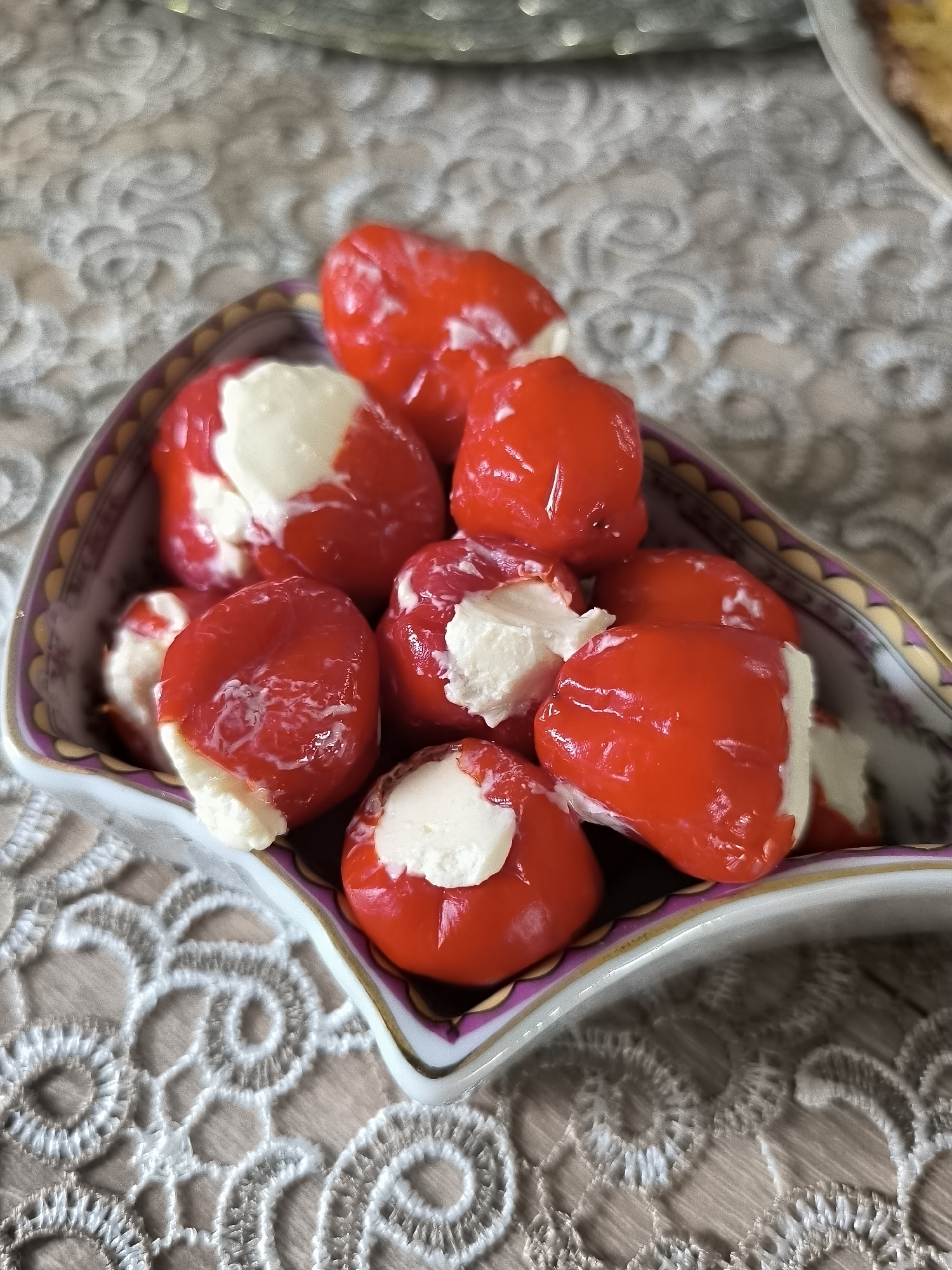 A heart-shaped or irregularly contoured serving dish, white with a purple and gold patterned rim, contains approximately 10 to 12 small, spherical to ovoid red peppers. Each pepper is visibly stuffed with an irregular mass of creamy white filling, which protrudes from an aperture in the pepper's skin. The peppers are glossy and appear moist. The dish rests on a light beige or off-white textile surface featuring an embossed or embroidered lace pattern with prominent swirling and floral motifs. The background is out of focus, showing indistinct shapes that suggest additional table settings or room elements. No individuals are present in the image. This scene is situated in the city of Dercekliai, Lithuania.
