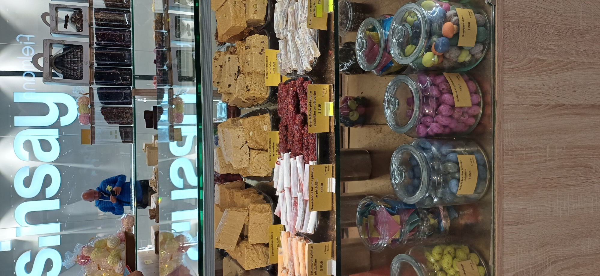 This is a medium shot, eye-level, indoor photograph of a Sinsay store's confectionery display. The store name, "Sinsay," is prominently displayed in large, illuminated blue letters across the top of the image. The display features various sweets, including halva in large blocks, rolled candies, and small, colorful candies in glass jars. A man in a blue hoodie is visible in the background, looking at his phone, reflected in the glass of the display case. The display is divided into sections, with yellow price tags indicating the cost and type of sweets. The lighting is bright and even, illuminating the products. The overall environment appears to be a retail setting, likely a department store or a dedicated confectionery shop within a larger complex, given the prominent branding.