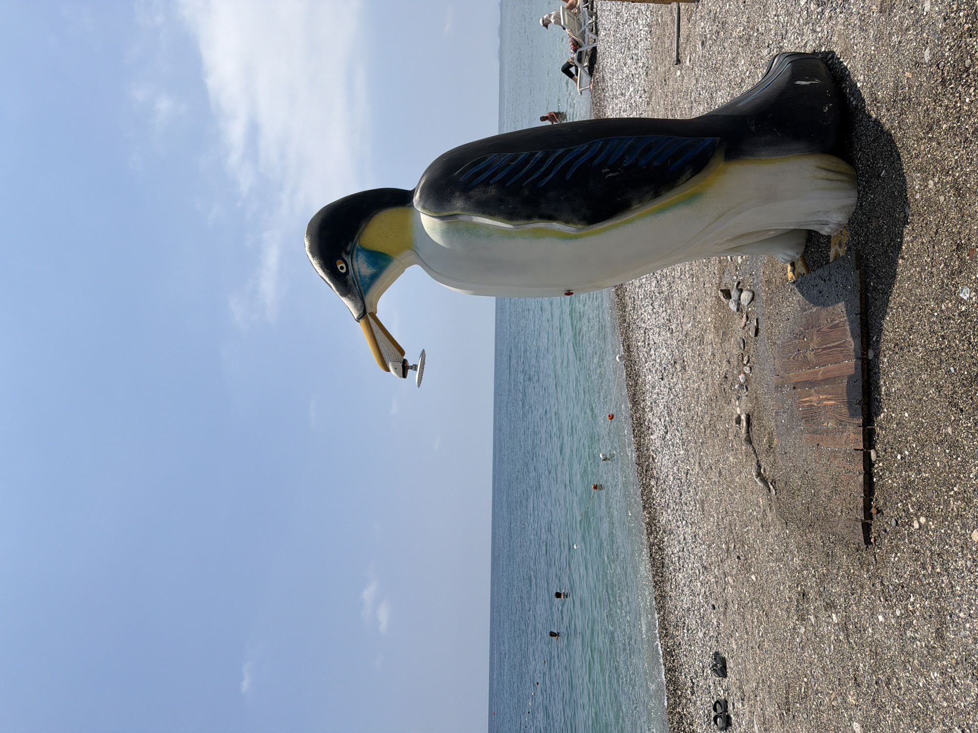 A life-sized, colorful statue of a penguin stands on a pebble beach in Kemer, Turkey, holding a fish in its beak. The penguin statue is predominantly white with black wings, a yellow chest, and blue accents around its neck and eyes. It is positioned facing the turquoise sea, which is calm and has a few people swimming in it. The beach is covered in small, gray pebbles, with some driftwood and a small wooden platform near the statue's feet. In the background, a few people are seen relaxing on lounge chairs, and a small buoy line is visible in the water. The sky is clear and blue with a few scattered white clouds. The overall scene depicts a sunny day at a beach resort.
