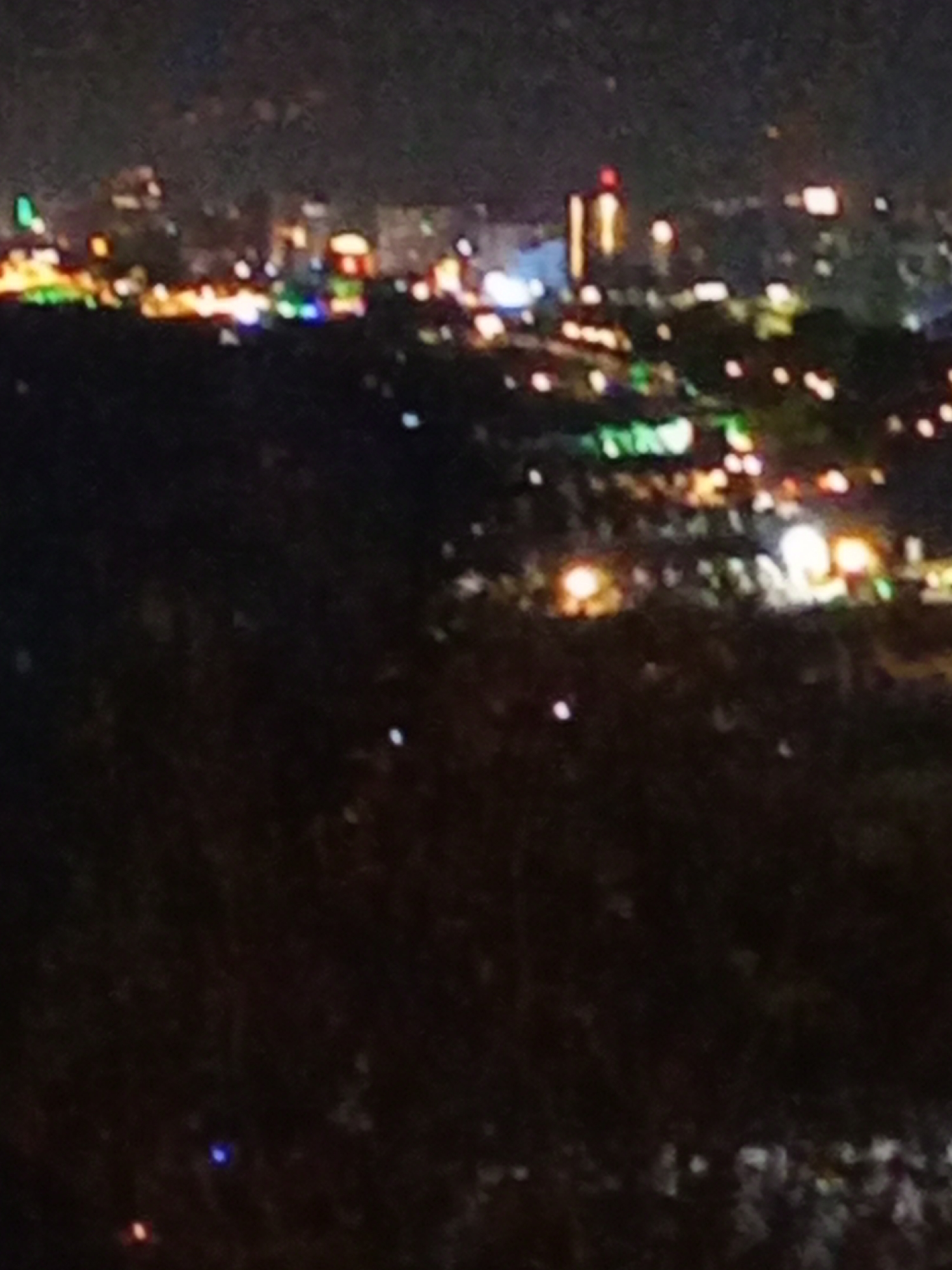 The image depicts a blurry nighttime cityscape, likely from an elevated viewpoint overlooking the city of Antalya, Turkey. The foreground is dark and dominated by indistinct shapes of trees or foliage. In the midground and background, numerous lights from buildings and streetlights create a diffused glow across the scene. Some of these lights appear in various colors, including white, yellow, green, and red, suggesting a vibrant urban environment. A tall, illuminated structure, possibly a building or tower, is visible in the distance, with a red light at its peak. The overall impression is of a sprawling city at night, with the lights creating a hazy and somewhat abstract pattern. The image quality is low, with significant blurriness and noise, making specific details difficult to discern. There is no clear indication of people or specific activities.