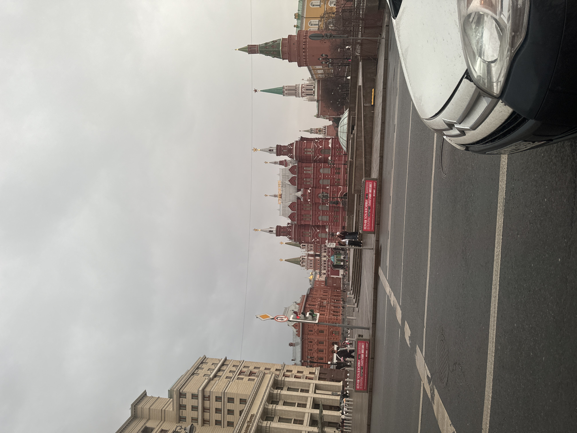 The image, taken on an overcast day in Moscow, Russia, features a view of historical landmarks. In the background, the red brick walls and distinctive towers of the Moscow Kremlin are prominent. The State Historical Museum stands out, its architecture reflecting its historical significance. To the left, the corner of a building, possibly the Four Seasons Hotel, is visible, adding a layer of contemporary urban life to the scene. In the foreground, a portion of a white car is present on the right. A road sign and pedestrian traffic light stand near the edge of the pavement, indicating a road with pedestrian crossings. A street with marked lanes is visible in the foreground. Several people are visible in the midground, walking or standing near the monuments. Signs in Russian can be seen by the side of the road, likely advertisements or informational markers.