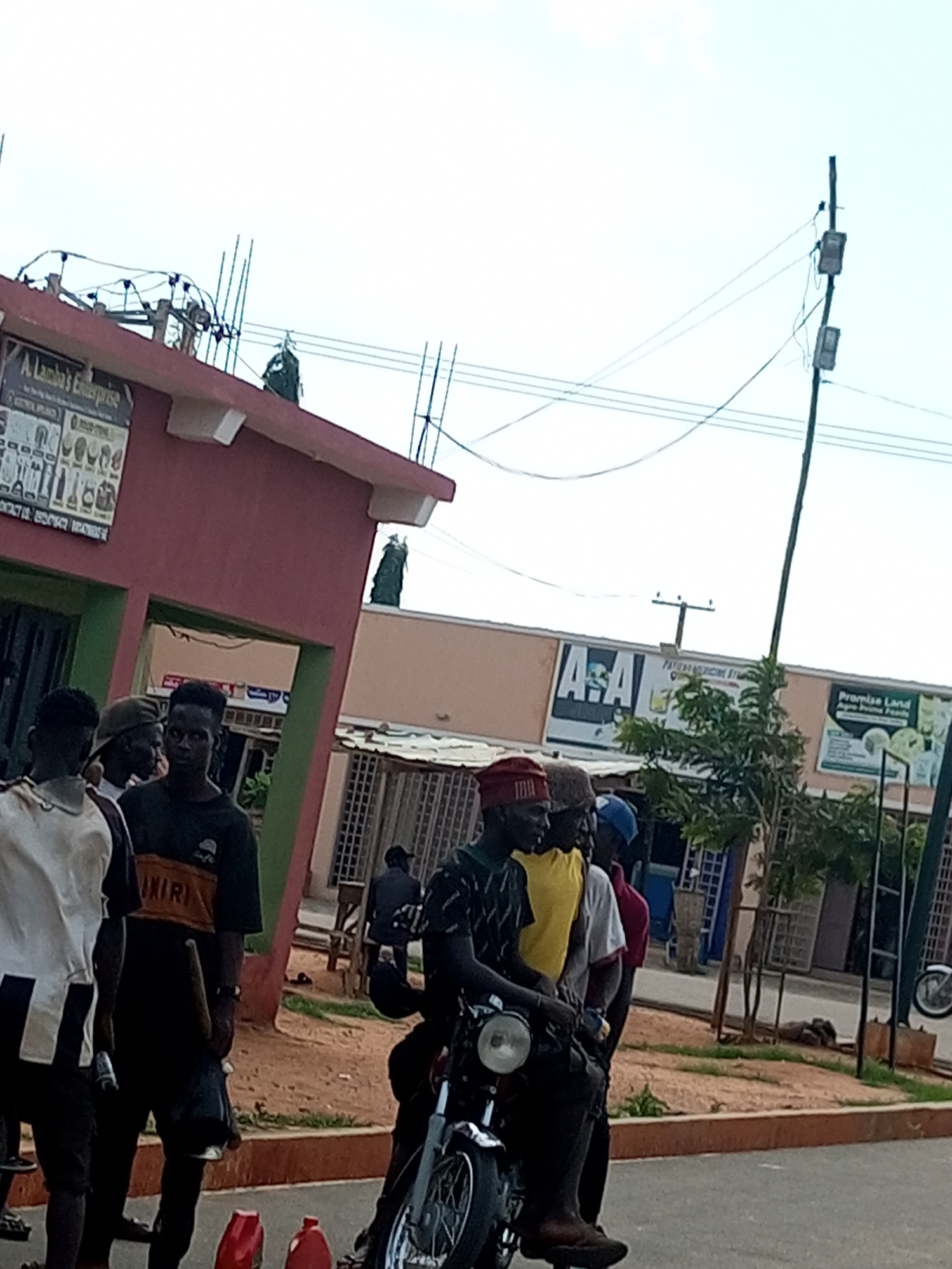 The image depicts an outdoor scene in Tempure, Nigeria, with a group of young men gathered on a street. In the foreground, four men are standing close together. The man on the far left wears a white and black shirt. Next to him, a man in a black t-shirt is partially visible. The third man from the left is wearing a yellow shirt. The fourth man is wearing a white t-shirt and a red cap. Behind them, a motorcycle is parked, with its front wheel and headlight visible.
Further back, a building with a pink facade is visible on the left, with a sign that reads "A. Lambo's Enterprise". To the right of this building, a longer, tan-colored building features signs for "A & A" and "Promise Land". Trees and electrical poles with wires are present in the background, indicating an urban or semi-urban setting. The ground appears to be a mix of dirt and pavement. Red plastic containers are placed on the ground in the lower foreground.