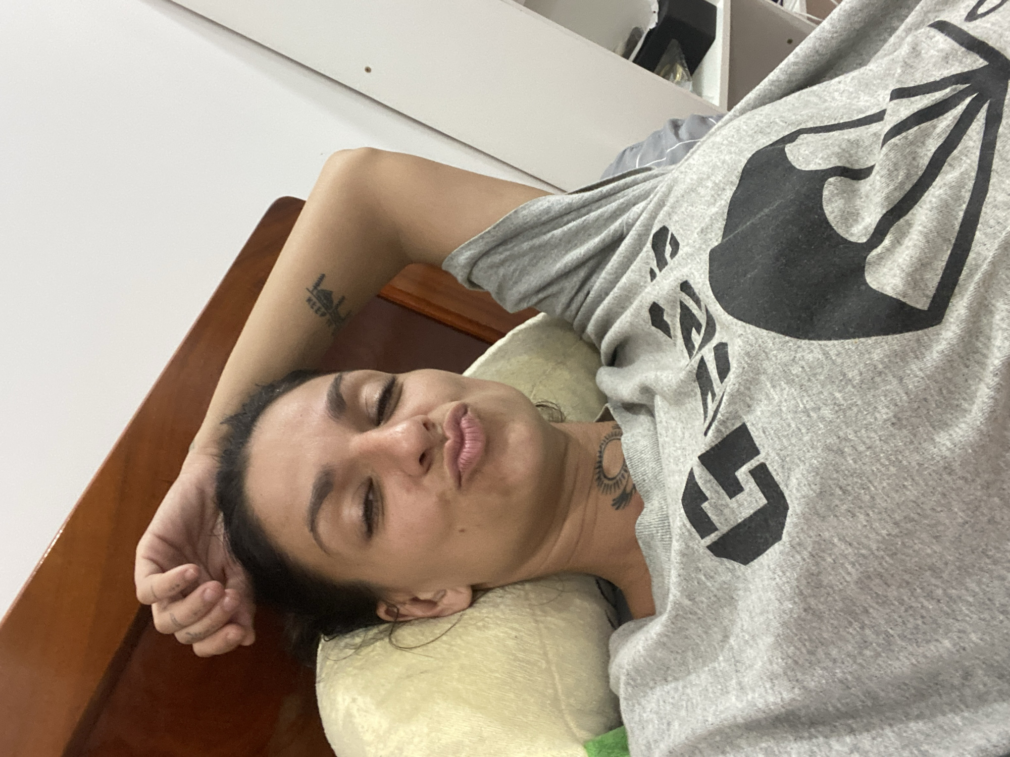 A woman is lying on a bed, holding her arm behind her head. She is wearing a grey t-shirt with a black graphic print and a tattoo on her neck. The room appears to be a bedroom with a wooden headboard and a white wall. The woman's facial expression is playful, with her eyes closed and her lips pursed. The lighting suggests it is daytime. The location is identified as Mũi Né, Vietnam.