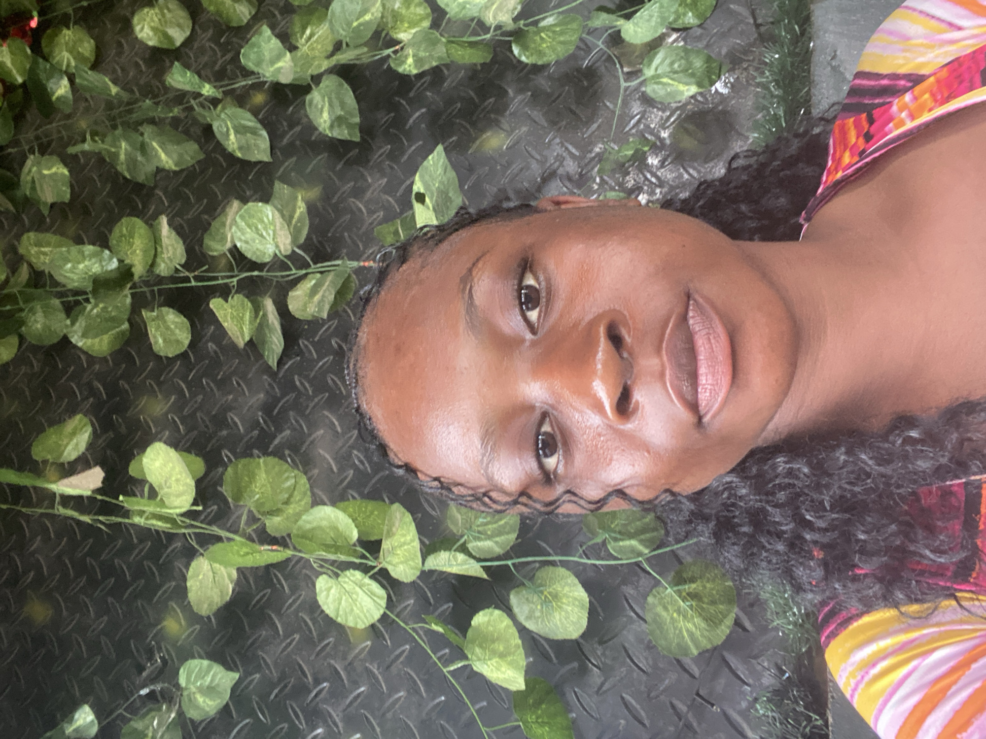 This media file is a close-up, portrait-style photograph of a woman lying down, with her head turned towards the camera. She appears to be outdoors, lying on a dark, textured surface that resembles diamond plate metal. Surrounding her are artificial green vines with heart-shaped leaves, creating a lush, jungle-like atmosphere. The lighting suggests it might be daytime, with no harsh shadows visible, indicating an overcast or diffused light source. The woman has dark skin, dark curly hair, and is wearing a brightly colored top with horizontal stripes in pink, yellow, orange, and white. Her expression is neutral, and her eyes are open, looking directly at the viewer. One of her eyes, the left one, is more prominently in focus. Her lips are slightly parted. The background is dark and out of focus, with the green vines and the metallic surface being the most distinct elements besides the woman herself. There is no visible text in the image. The overall mood is serene and somewhat artistic, with the vibrant colors of her clothing contrasting with the darker background and greenery. The setting could be interpreted as a creative photoshoot or a casual pose in a garden-like environment. The location context provided is Jalingo, Nigeria, which does not offer any specific visual cues within the image itself, but provides an additional layer of information about the origin of the media.