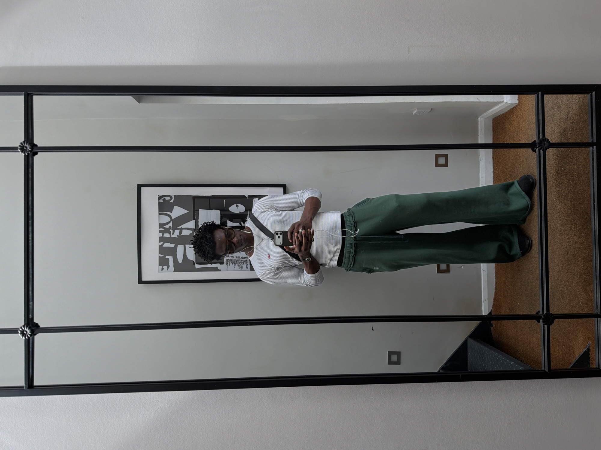 A person with dark skin is reflected in a rectangular, black-framed mirror. The person wears eyeglasses, a striped long-sleeved shirt, green pants, and dark shoes. They are holding a smartphone and wearing headphones. Behind the person in the reflection, there is a framed black and white art print on the wall. The reflection reveals an adjacent room with a light-colored carpet and two electrical outlets on the wall. The mirror frame has decorative floral details at the intersections of the frame elements. The location is London, United Kingdom.