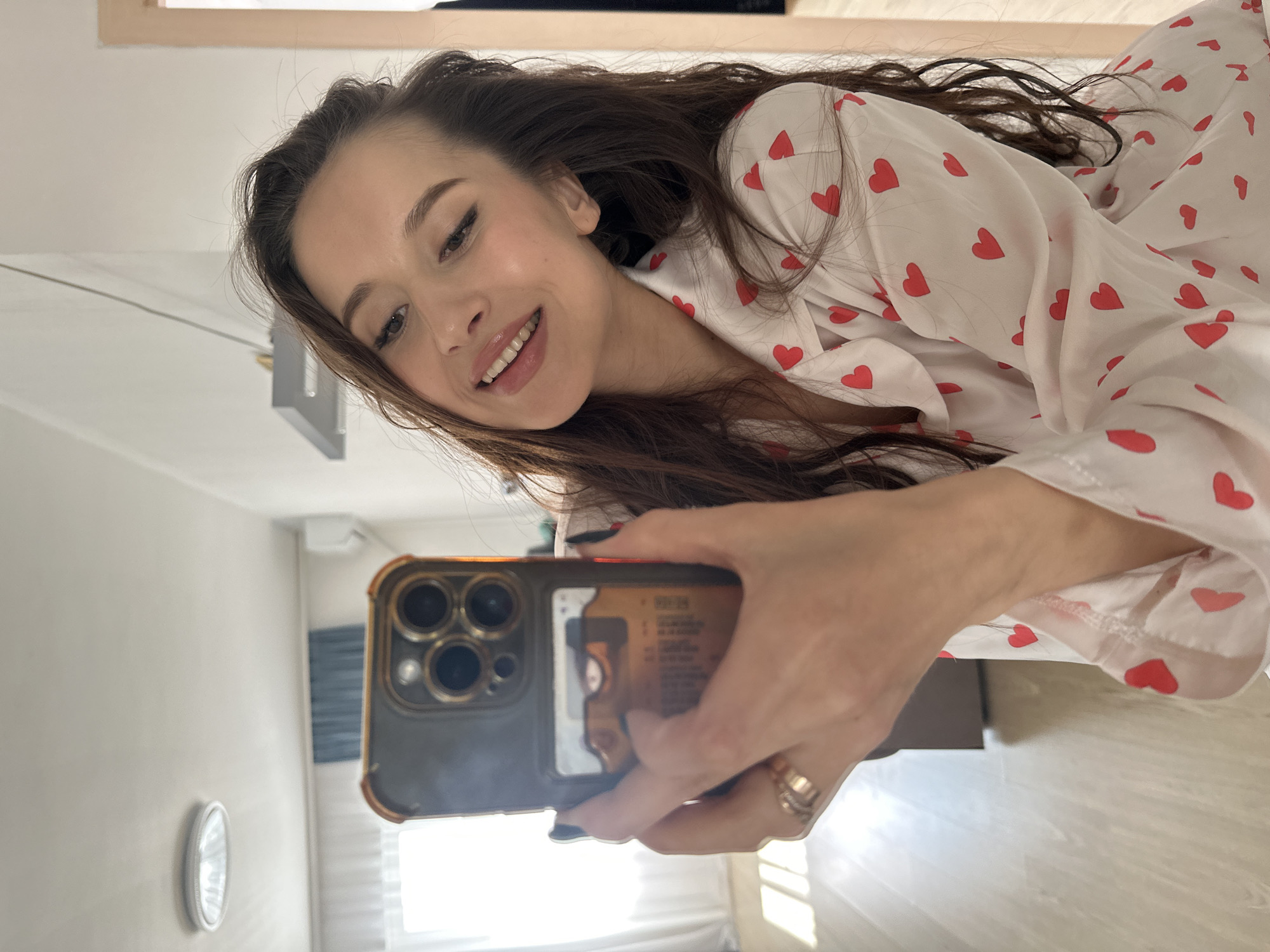 A woman is taking a selfie in what appears to be a room with light-colored walls and flooring. The woman has long brown hair and is wearing a light-colored pajama top with red hearts. She is holding a cell phone with a dark case that has a gold or bronze-colored trim. The background shows elements of a room including a window with light shining through, a wall with a round light fixture, and glimpses of furniture or fixtures. No specific details indicate the location is Yekaterinburg, Russia.