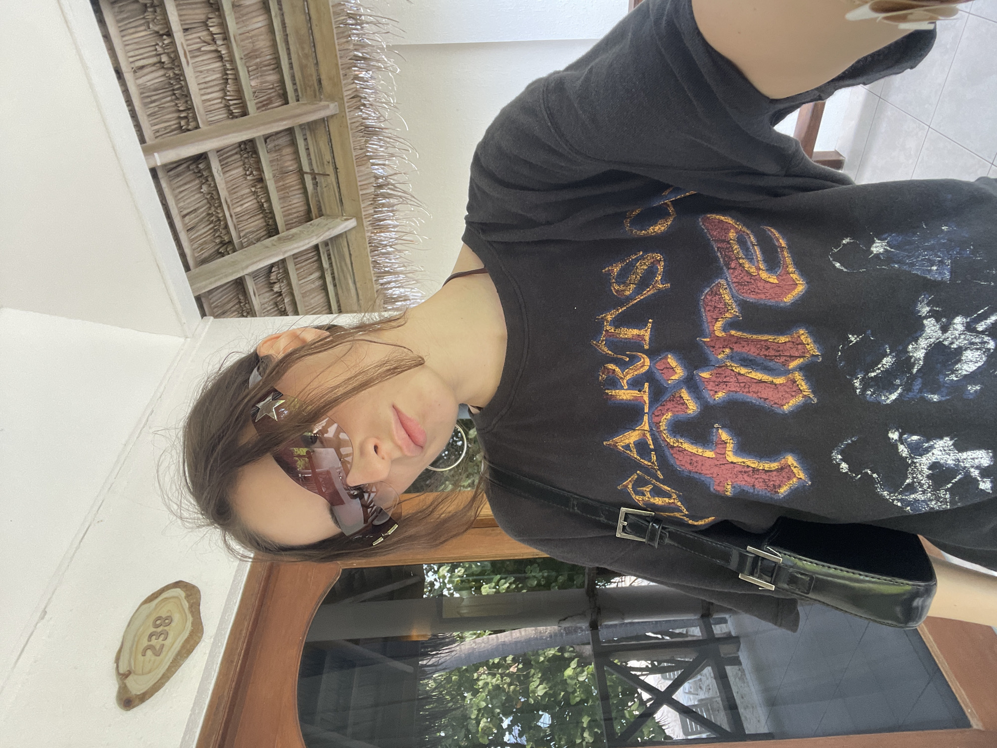 A young woman is taking a selfie in a tropical location. She is wearing oversized sunglasses with a futuristic design, a dark t-shirt with a graphic print, and a small black shoulder bag. The background shows a thatched roof structure, indicating an outdoor setting. A wooden sign with the number "238" is visible on the wall above her. To the left, a glass-enclosed porch with wooden railings and lush greenery can be seen, suggesting a resort or hotel environment in the Maldives. The lighting indicates it is daytime.