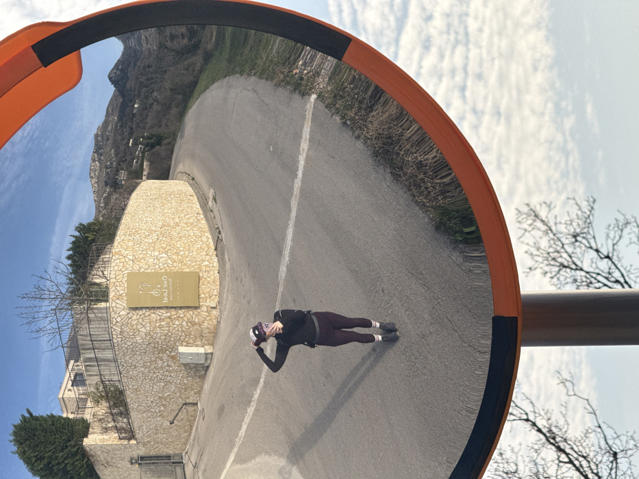 A convex traffic mirror with an orange and black frame reflects a outdoor scene. The central subject is a female individual standing on a paved asphalt road, directly facing the mirror. She is wearing a dark long-sleeved top, dark leggings, dark shoes, and white socks with two horizontal stripes. A small dark backpack is visible on her back. Her right hand holds a mobile phone, and her left hand is raised to her head, adjusting a dark baseball cap/visor. Her elongated shadow is cast to her left on the road surface.
Behind the individual, the paved road curves sharply to the right and ascends. A single white line is painted horizontally across the road. To the left of the road, a substantial retaining wall constructed from light-colored, irregularly shaped stones supports a building that is partially visible behind it. A metal gate is present on the far left. Affixed to the stone wall is a rectangular, light-brown sign with inverted text, clearly legible as "Oak Leaf" with stylized tree/leaf designs, and "Restaurant" below it. This establishment is located in Vojnići, Montenegro. Beyond the curving road and structures, distant mountains covered with sparse vegetation rise under a clear blue sky punctuated by scattered white clouds. The lower portion of the mirror reflection reveals a segment of a tree with bare branches against the sky. The scene is illuminated by direct sunlight, indicated by the distinct shadow of the person.
