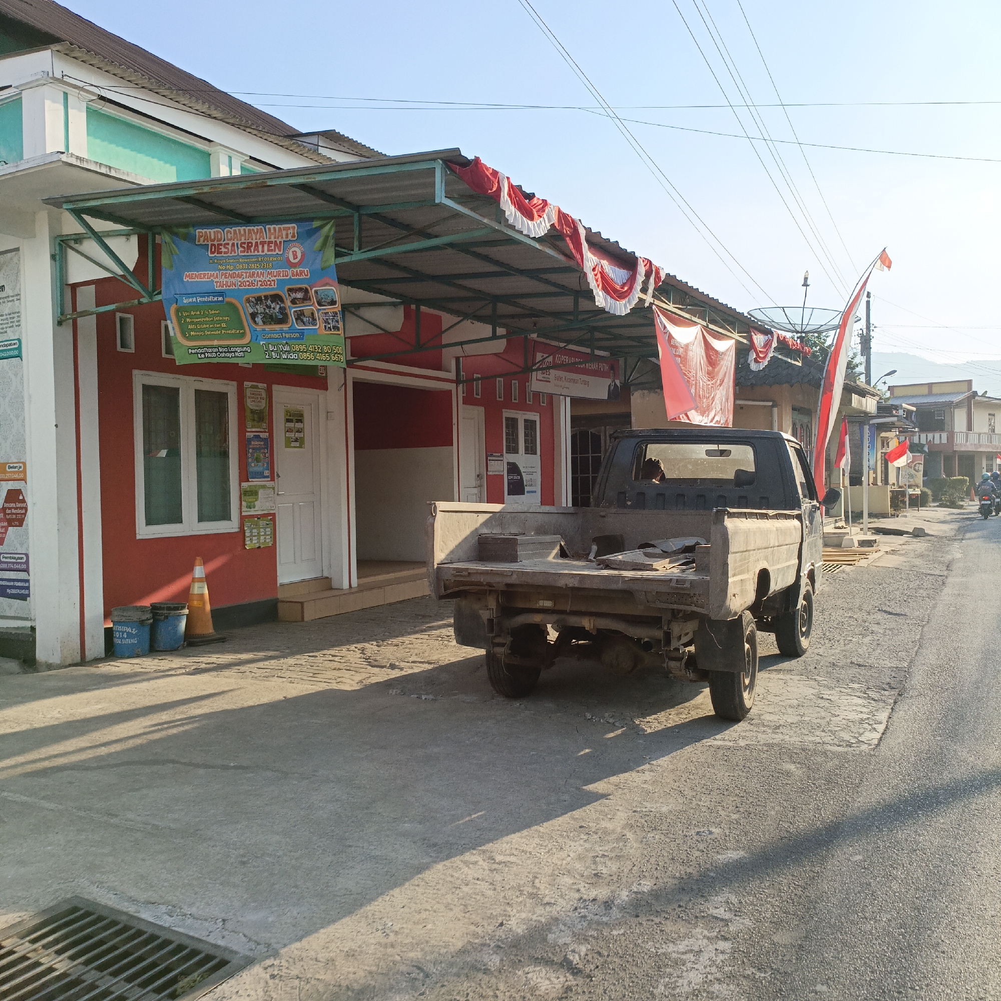 This is a street-level, full-shot image taken in Sraten, Indonesia. The scene depicts a narrow asphalt road with a concrete sidewalk. In the foreground, to the left, is a building with a red facade and white trim, featuring two windows and a door. A banner for "PAUD CAHAYA HATI DESA SRATEN" is displayed on the awning. Two blue buckets and an orange traffic cone are on the sidewalk near the building. To the right of the building, a dark-colored pickup truck is parked on the road, facing away from the camera. The truck bed contains some debris. Further down the road, there are other buildings, more Indonesian flags and banners, and a satellite dish on a pole. The sky is clear and blue, and the sun casts long shadows across the road and sidewalk. The overall impression is that of a community setting in a rural or semi-urban area of Indonesia.