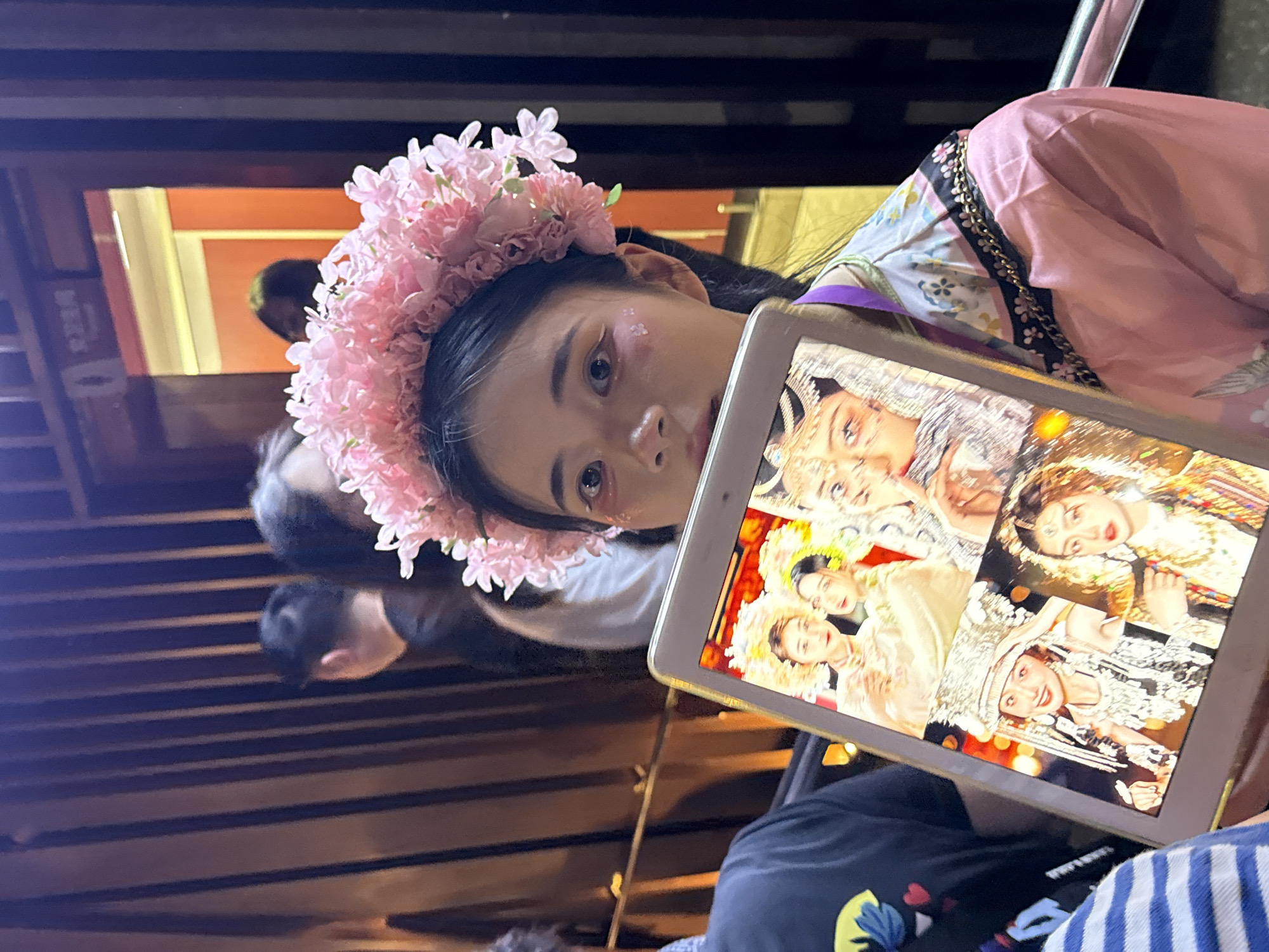 A young woman is prominently featured in a dimly lit indoor setting, captured from a slightly high angle, looking directly towards the camera. She wears a large, ornate flower crown made of pink blossoms on her dark hair. Her face is made up, with subtle glitter or small flower-shaped appliques near her left eye, and she has a calm, focused expression. She is wearing what appears to be traditional or themed clothing; a pink garment with a patterned sleeve featuring floral designs and a gold chain detail is visible on her right side.
In her hands, she holds a tablet, angled towards the viewer, displaying a collage of several images. These images show different individuals, primarily women, dressed in elaborate traditional costumes, rich in color and intricate patterns, with detailed headwear and jewelry, suggesting cultural or theatrical attire.
The background is dark and appears to be a wooden interior, possibly a restaurant, performance venue, or a themed experience. Blurry figures of other people are visible in the background, suggesting a public or semi-public space. One figure directly behind the main subject is also blurred. The ambient lighting is warm, possibly from artificial sources, casting a soft glow on parts of the scene. The overall impression is that of a posed moment, perhaps during an event or gathering, in Yuzhong District, China, where the woman is showcasing images on her tablet while also acknowledging the camera. There is no clearly legible text in the image.