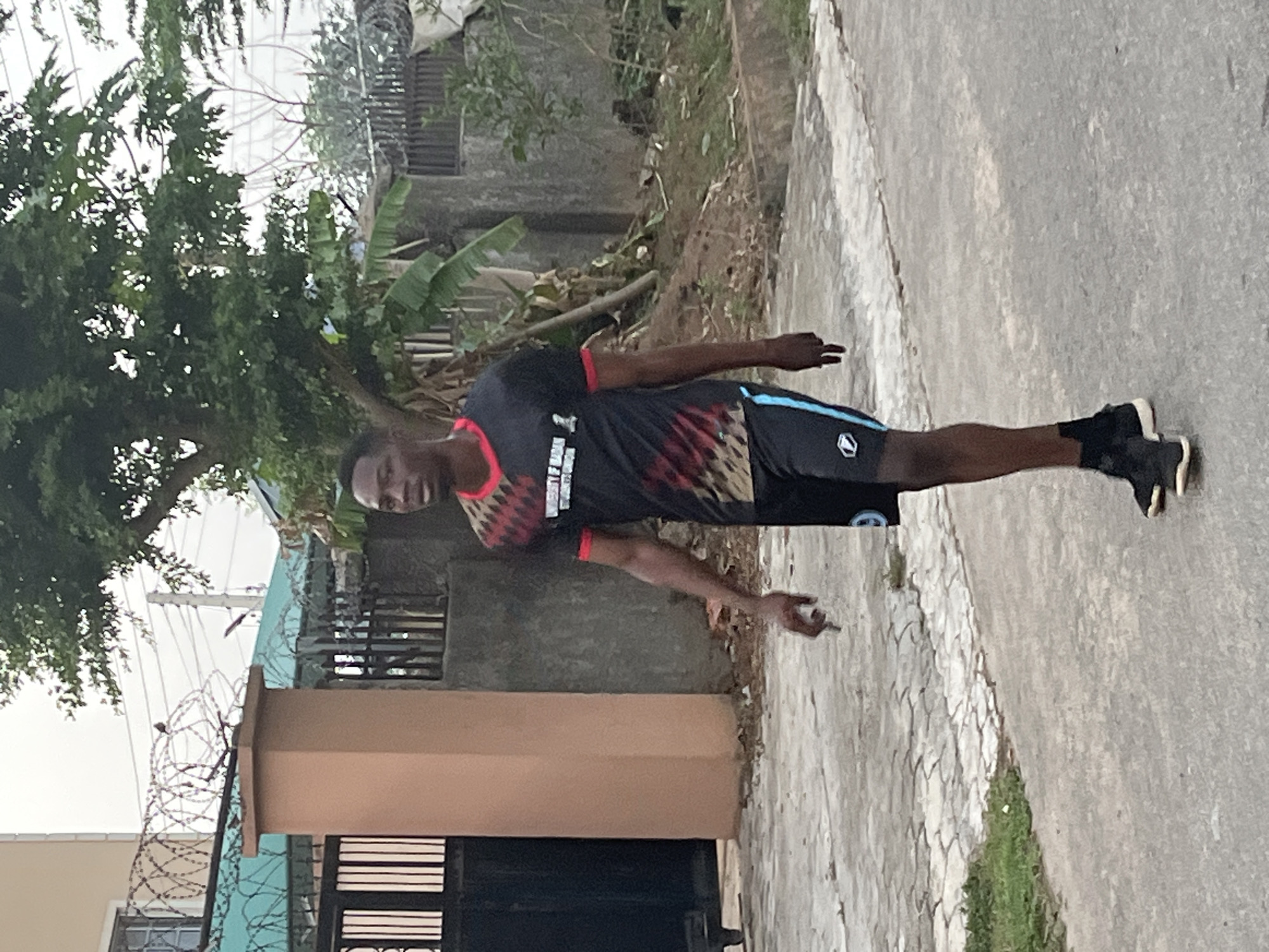 A person stands on a paved sidewalk in what appears to be a residential area in Abuja, Nigeria. The individual is wearing a black and red patterned sports shirt, black shorts with a blue stripe, and black sneakers. He is facing the camera with his arms extended.
Behind the person, there's a building with a gate secured by barbed wire. The surrounding environment includes greenery, such as trees and vegetation, and concrete structures like walls and fences, which suggest a mix of residential and developed areas. The sidewalk shows a pattern of interlocking pavers.