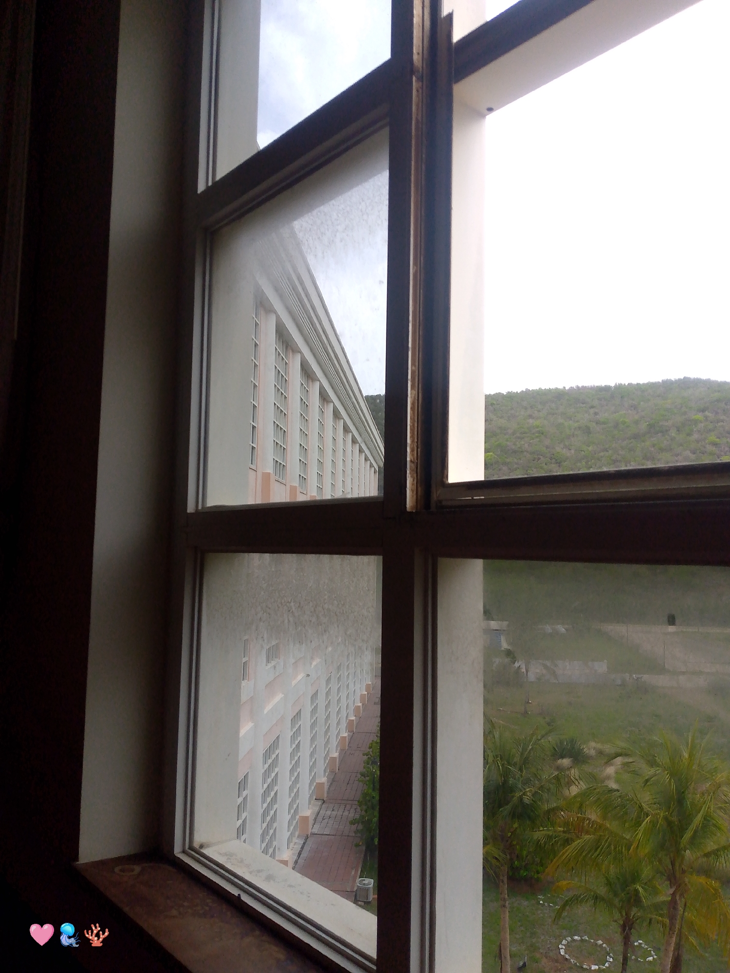 This media file is a photograph taken from inside a building, looking out through a window. The window frames a view of a long, multi-story building with many windows, likely a hotel or residential complex, extending into the distance. The building appears to be light-colored with pink accents.
Beyond the building, a green, tree-covered hillside is visible under a bright, overcast sky, suggesting it is daytime. The weather appears to be cloudy or possibly hazy. The foreground shows a wooden windowsill with some faint smudges and a pink heart, a blue jellyfish, and a coral reef emoji, adding a touch of digital embellishment to the scene. The window itself has a slightly grimy appearance, with streaks and dust visible on the glass.
The overall setting suggests a tropical or sub-tropical location, given the presence of palm trees visible in the lower right portion of the frame. The atmosphere is somewhat muted, perhaps due to the overcast weather, and the scene evokes a sense of looking out from a temporary dwelling or accommodation. The building's architecture hints at a resort or a large complex designed for many occupants. There is no discernible text in the image.