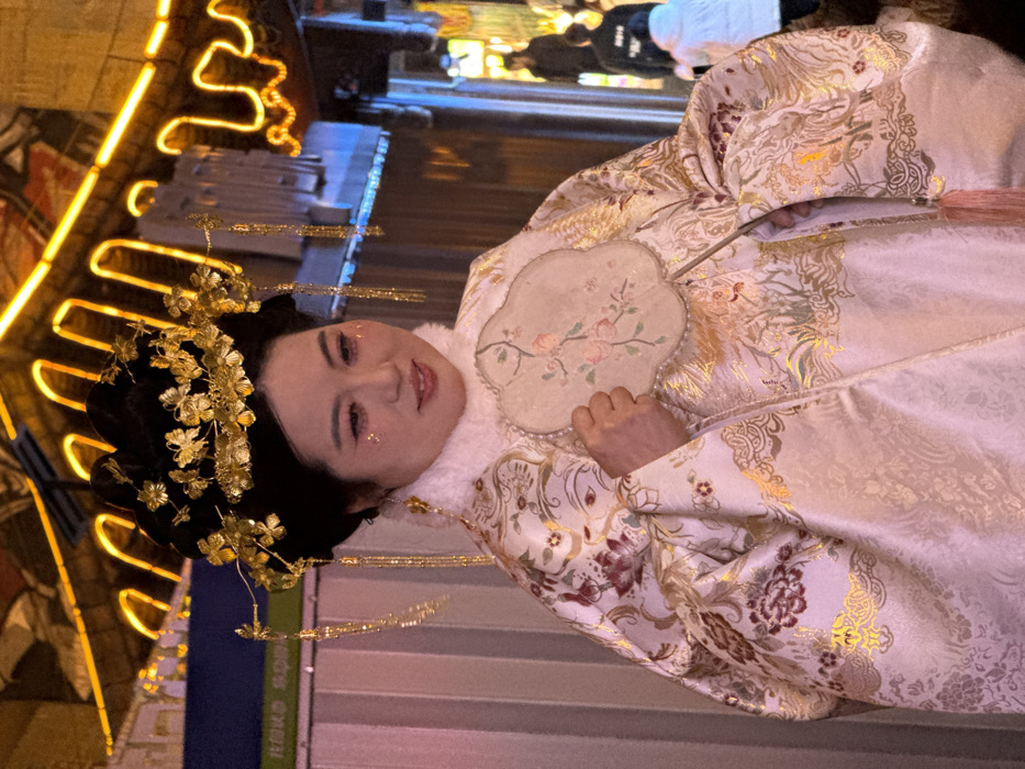 The image depicts an East Asian female positioned centrally within an illuminated nighttime street in Yuzhong District, China. She is slightly turned to her left, with her head angled downwards and to the right, displaying a gentle smile. Her dark hair is styled in an elaborate updo, heavily adorned with gold-colored metal ornaments resembling flowers and leaves, complemented by golden tassels dangling on each side. Her facial makeup includes pink eyeshadow, eyeliner, and iridescent glitter applied beneath her eyes. She wears a cream or off-white brocade outer garment, likely a robe or coat, featuring a white fur collar. This garment is richly patterned with gold, burgundy, and other muted colors, depicting floral and intricate decorative motifs. In her right hand, she holds a scalloped, rigid fan-shaped object, possibly made of silk or paper, bordered with small pearls or beads, and adorned with a painted motif of pink and green flowers. The background consists of a street environment illuminated by vibrant orange, yellow, and blue neon lights that outline traditional Chinese architectural elements and rooflines. Multiple blurred figures are visible in the distant background to the right, suggesting a busy public thoroughfare.