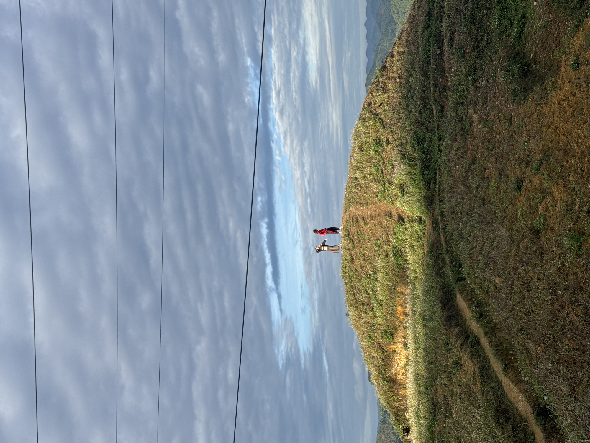 Two people stand on a grassy hillside in Lũng Cú Commune, Vietnam. One person wears a red top. Power lines cross the sky. The sky is cloudy. A path runs along the hillside.
