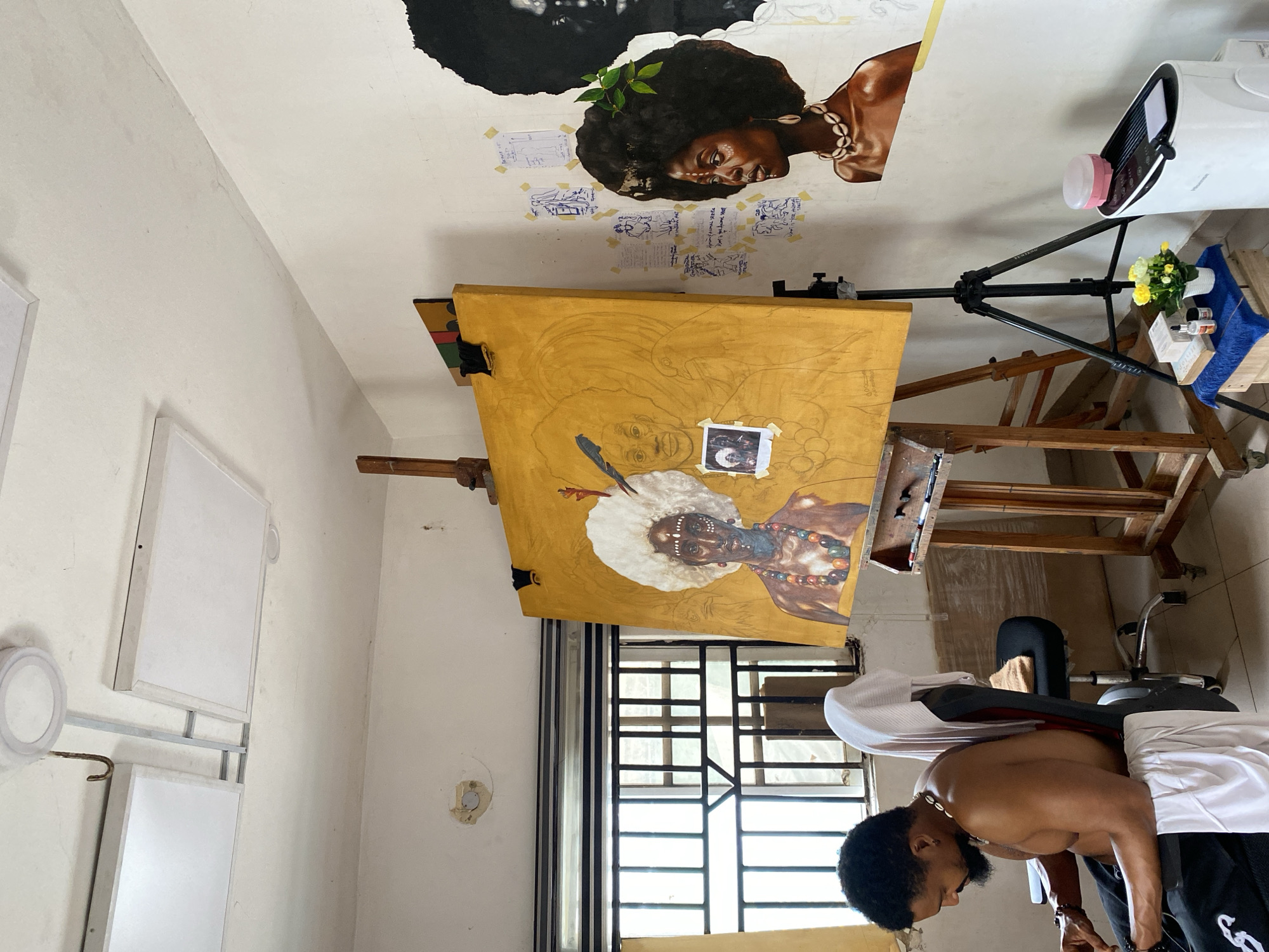 A man is bent over, presumably working on an art piece in a room in Ado, Nigeria. The room features art on the ceiling and an easel holding a yellow canvas with a partially completed painting of a figure wearing a feathered headdress. A projector is set up on a tripod stand. There are also art panels mounted to the wall, and a barred window. The man himself has dark hair and a beard, is shirtless, and appears to be wearing a white shirt and black pants.