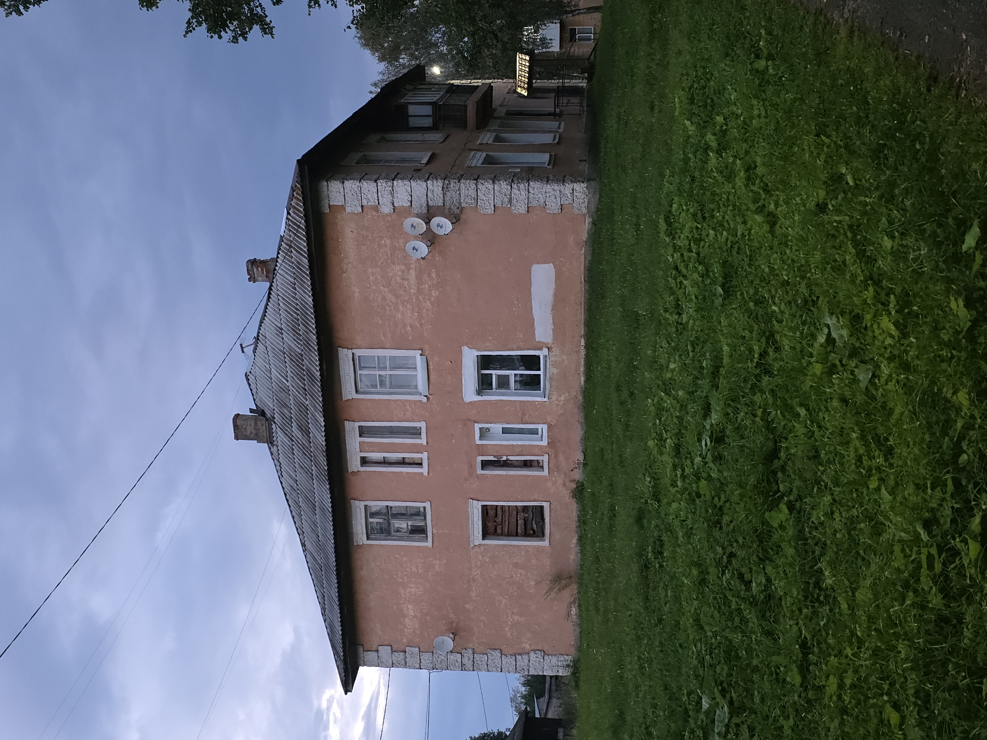An exterior, eye-level, full shot shows a weathered, two-story apartment building with a pale orange facade and a gray, corrugated roof. The building is situated in the city of Magnitka, Russia. The structure has a stone border around the corners and is adorned with several satellite dishes on its front and side. Multiple windows are visible, some with white frames, while one on the lower left is boarded up with wooden planks. A small balcony with a railing extends from the second floor on the right side of the building. In front of the building is a lush, green lawn that slopes gently upwards. The sky above is overcast and a pale blue-gray, with visible power lines stretching across the frame. To the right of the building, trees with green foliage are present, and a pathway runs along the edge of the lawn. The overall scene suggests an early evening setting in a residential area.