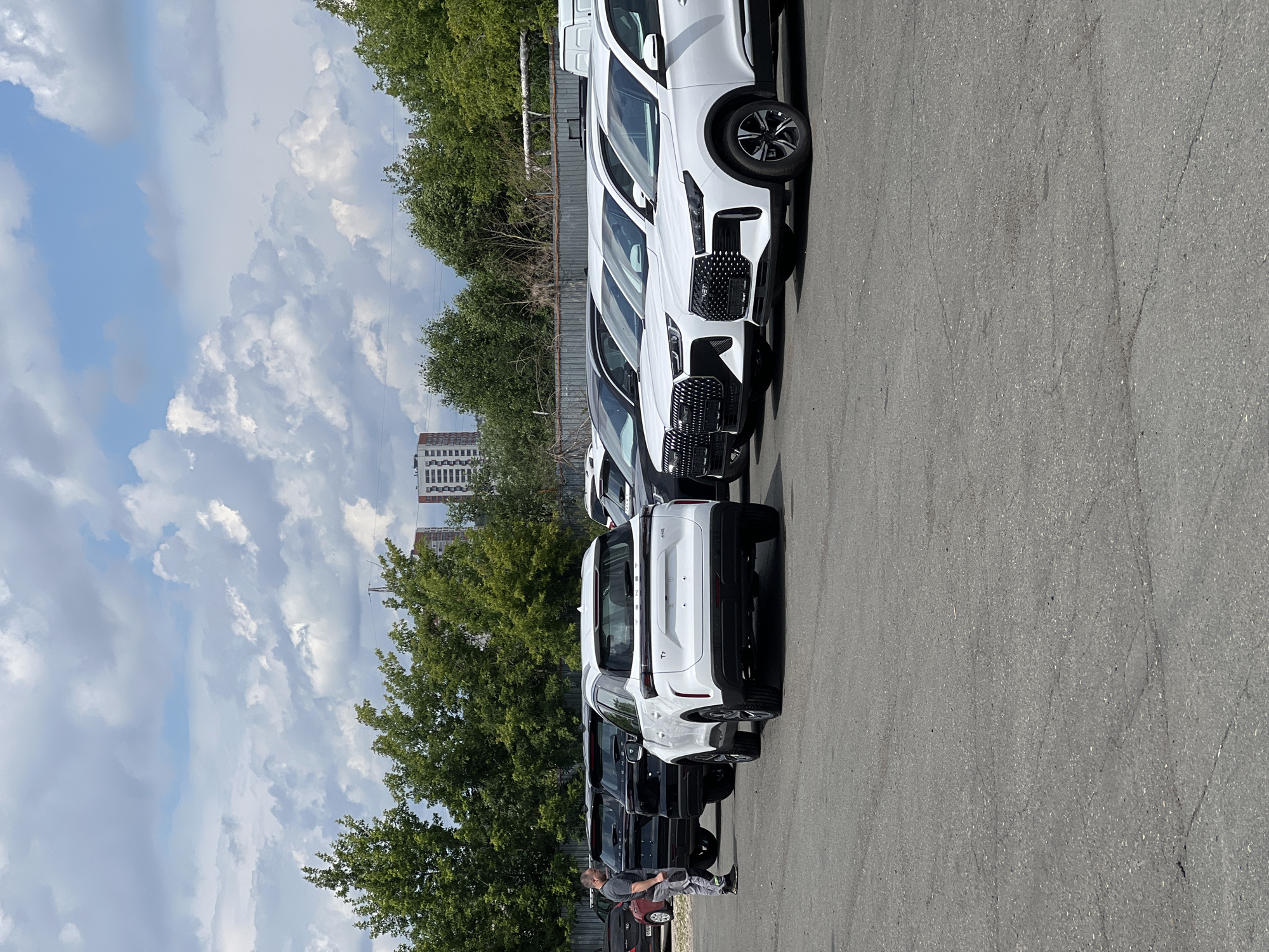 A group of white and black SUVs are parked in an outdoor lot. A man in a grey t-shirt and pants stands near a white SUV, looking at it. Buildings and trees are visible in the background under a cloudy sky. The scene is likely a car dealership or a parking lot in Izhevsk, Russia, given the presence of the brand "TENET" on the vehicles, which is a Russian automotive brand.