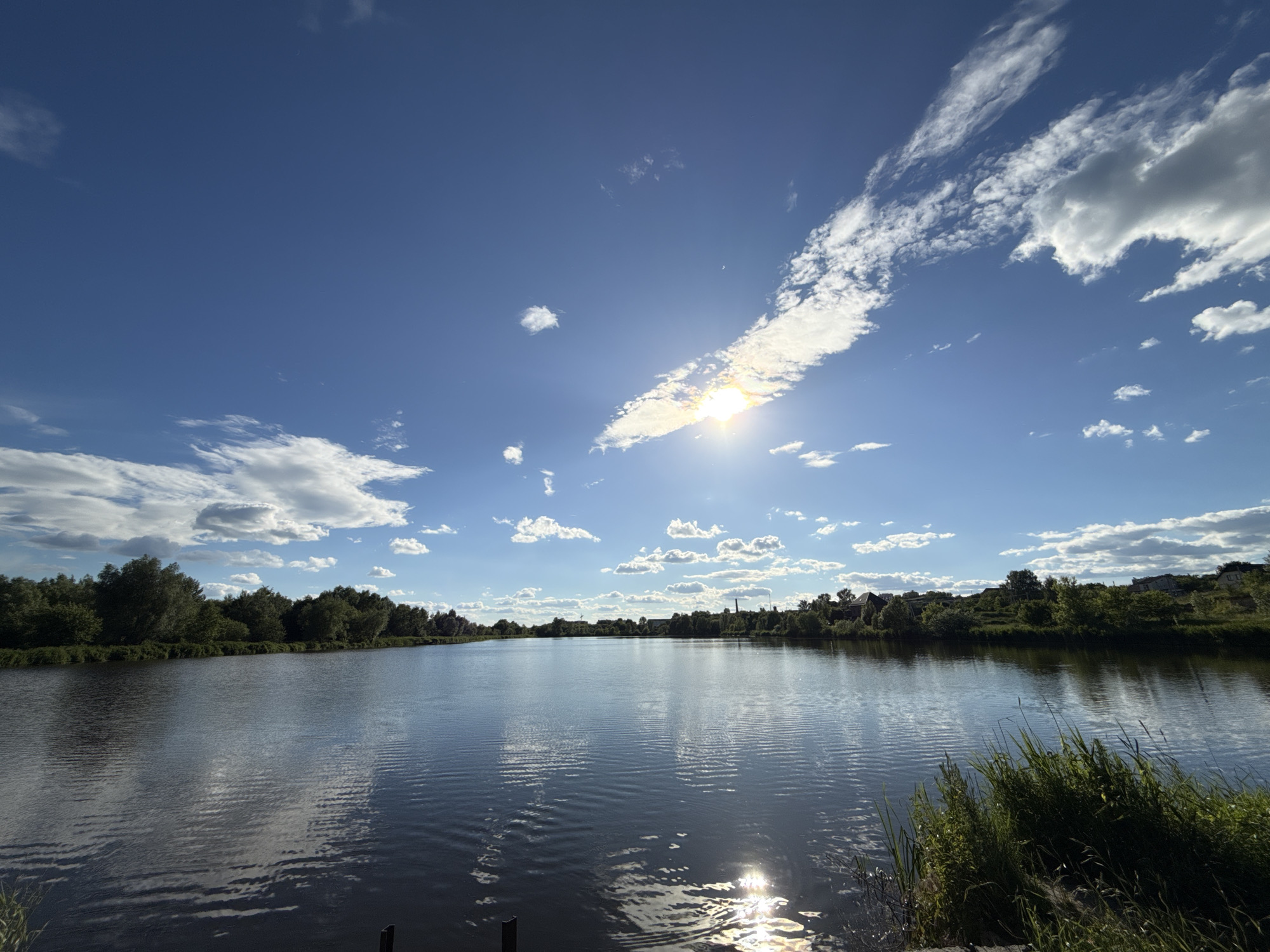 A wide, tranquil river reflects the bright blue sky and scattered clouds. The sun shines intensely, creating a glare on the water's surface. The riverbanks are lined with lush green trees and vegetation. In the distance, on the far bank, a few buildings and a church steeple are visible, suggesting the presence of a settlement. The overall scene depicts a peaceful, natural landscape with a hint of civilization. The location is the city of Hostomel, Ukraine.