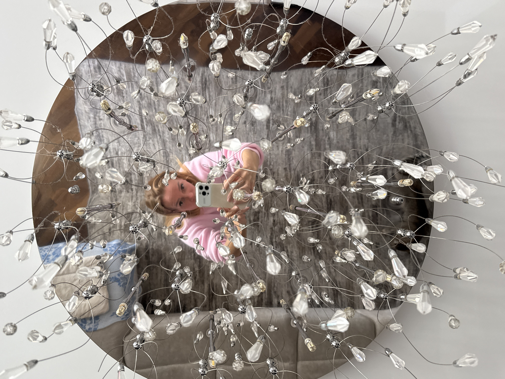 A reflection in a round mirror reveals a woman taking a selfie in an interior setting. The mirror is adorned with a chandelier-like fixture featuring numerous clear crystal or glass elements connected by thin, silvery metal rods.
The reflected room contains a bed covered with a patterned grey bedspread, and a blue-patterned pillow is visible at the edge of the bed. A grey sofa or upholstered bench sits nearby.
The woman in the reflection wears a pink top and holds a smartphone to capture the photo. Her blonde hair is styled loosely. The setting suggests a contemporary or modern apartment. There's no specific evidence to link this scene directly to Saint Petersburg, Russia.