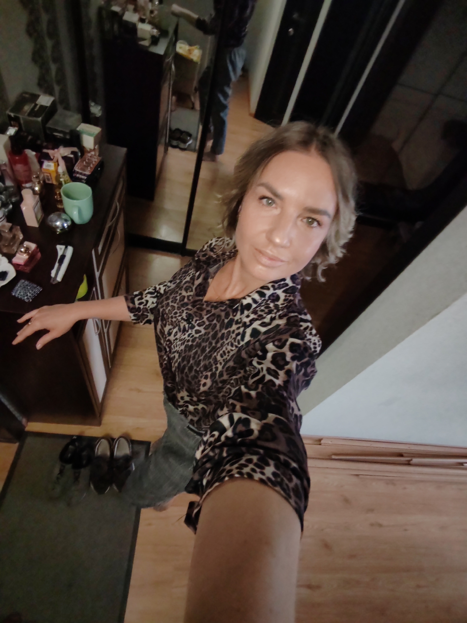 This full-shot selfie captures a woman indoors, likely in a home setting. The woman, with short, wavy light brown hair and light eyes, is wearing a dark brown and beige leopard print blouse and grey jeans. She holds the camera at arm's length, angled downwards, and looks directly at the lens with a neutral, composed expression.
To her left, a dark wooden dresser or cabinet is visible, topped with an array of personal items. These include several perfume bottles (some clear, some pink, one spherical silver), a light blue/green mug, small boxes, and other toiletries. Below the dresser, on a dark mat, are several pairs of dark shoes.
The background on the right features a large mirror reflecting parts of the room, including light wooden flooring and the lower body of a person (likely the photographer's reflection or another individual). The visible floor throughout the image is light-colored wood laminate. The lighting suggests a bright indoor environment. There is no clearly readable text on any of the objects in the image. The provided context indicates the location as Petropavlovsk-Kamchatsky, Russia, though this is not visually discernible within the photograph.