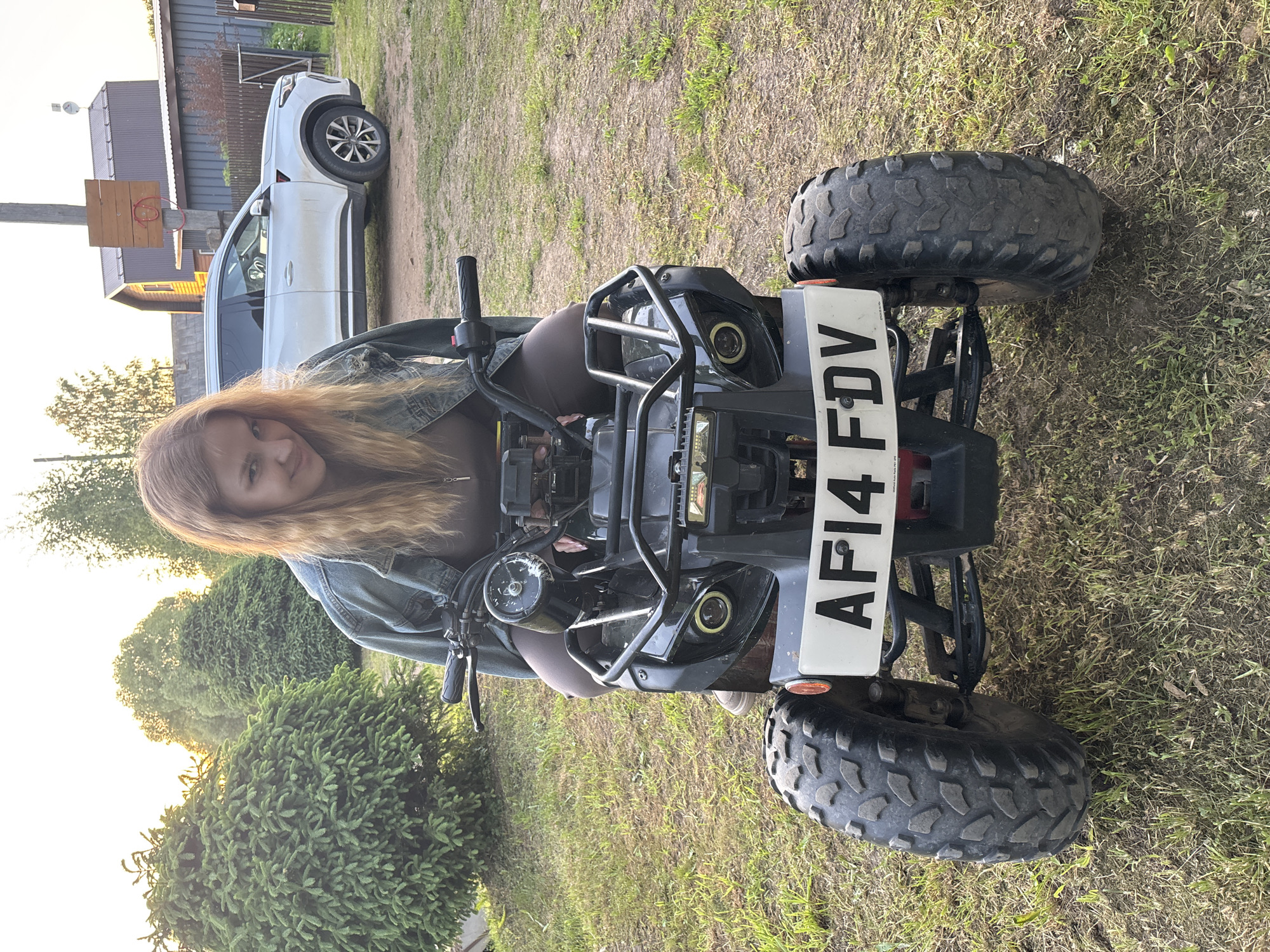 The image depicts an outdoor scene in what is identified as Rozhdestveno, Russia, featuring a woman seated on a quad bike. The woman, positioned centrally, has light brown hair and is wearing a blue denim jacket over a brown top and dark leggings, with light-colored footwear partially visible. She is astride a dark-colored quad bike, looking directly towards the camera with a slight smile. The quad bike has robust, knobbly tires, and a white front license plate displaying "AF14 FDV" is clearly visible. Its front includes a metal bumper guard and two distinct headlights, one round and one rectangular.
The setting appears to be a rural or semi-rural yard. To the left of the woman and quad bike, dense green evergreen shrubs and trees are visible, with bright sunlight emanating from behind them. To the right, a white SUV is parked on an unpaved surface. Behind the SUV, a wooden fence separates the area from a structure featuring a makeshift wooden basketball hoop attached to a utility pole. In the far background, two buildings are discernible: one a light-colored wooden house with a dark roof, and another, smaller building with a grey corrugated roof. The ground throughout the foreground and midground is a mix of dry grass and bare dirt. The lighting suggests either late afternoon or early evening.