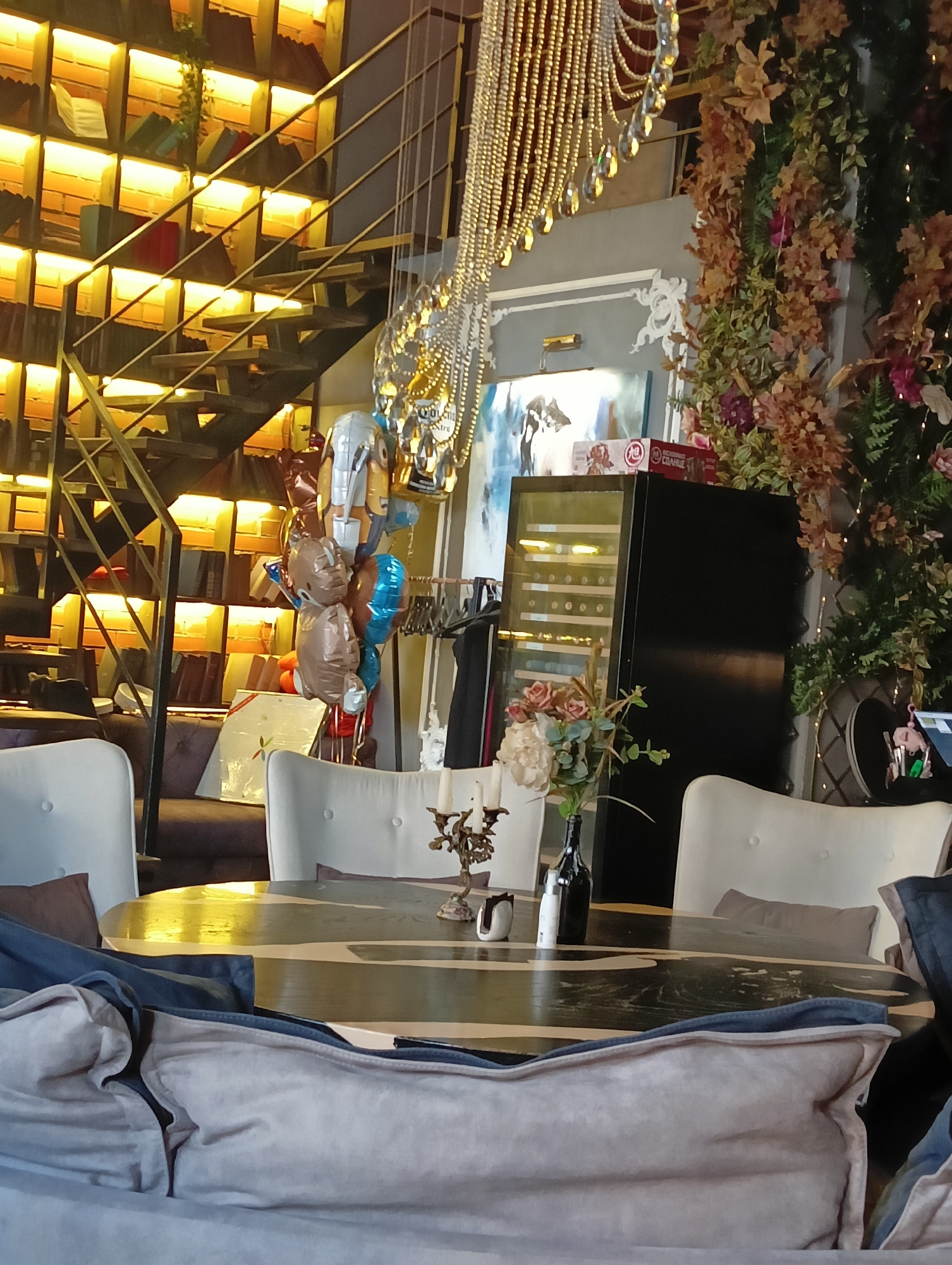 The image depicts an interior scene that appears to be a restaurant or cafe, featuring a blend of modern and rustic decor. A prominent element is a multi-tiered staircase with metal railings, situated in front of bookshelves illuminated from within by warm lighting. The bookshelves are filled with books and some decorative items, creating a cozy atmosphere.
To the right of the staircase, a large, ornate chandelier hangs from the ceiling. Adjacent to this, a lush display of artificial foliage and flowers cascades down, interspersed with fairy lights, adding a touch of nature and festivity.
In the foreground, a large, round dining table with a dark, patterned surface is visible. It is surrounded by several chairs, some of which are upholstered in white. On the table, a vase with a floral arrangement, a dark bottle, and other decorative items are placed. The lower part of the image shows plush seating, possibly beanbag chairs or sofas, covered in light-colored fabric.
Further into the background, a black wine cooler with glass doors reveals rows of wine bottles. Above the wine cooler, a framed piece of abstract art is mounted on the wall. A collection of balloons, some shaped like cartoon characters, are gathered near the staircase, suggesting a celebratory event. The overall color palette is warm, with dominant tones of brown, gold, and white, accented by the greenery and floral elements. The lighting is soft and ambient, enhancing the inviting ambiance of the space.