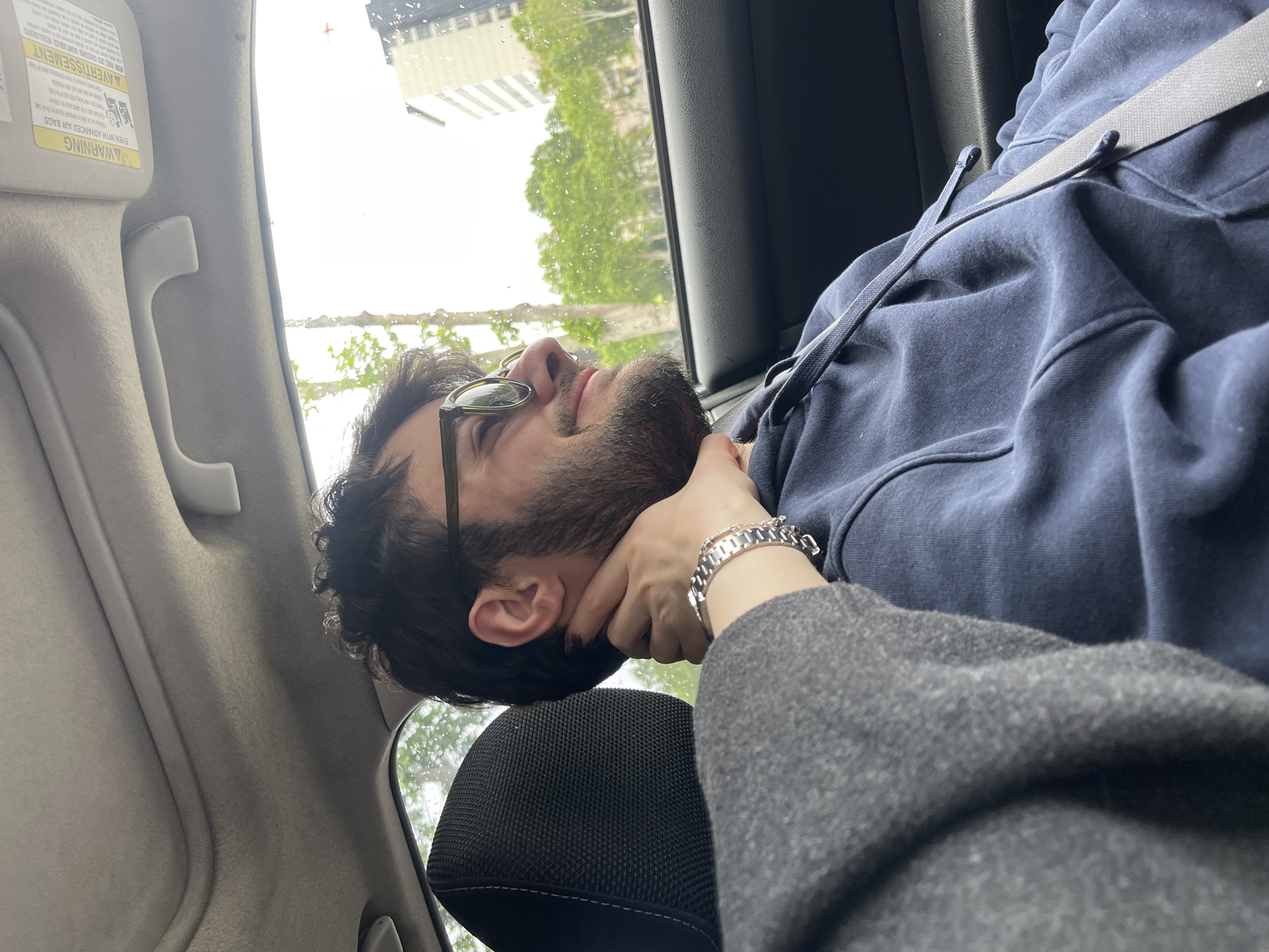 A person with dark hair and a beard is lying down in a car, possibly asleep. The person is wearing glasses, a navy blue hoodie, and a seatbelt. Another person's hand, adorned with a silver bracelet, is gently supporting the first person's head. The car window shows an out-of-focus view of buildings and trees, likely in an urban setting. Based on the visible architecture and general ambiance, the city is likely Tbilisi, Georgia. The interior of the car includes a gray headrest and a warning label on the sun visor. Raindrops are visible on the car window.