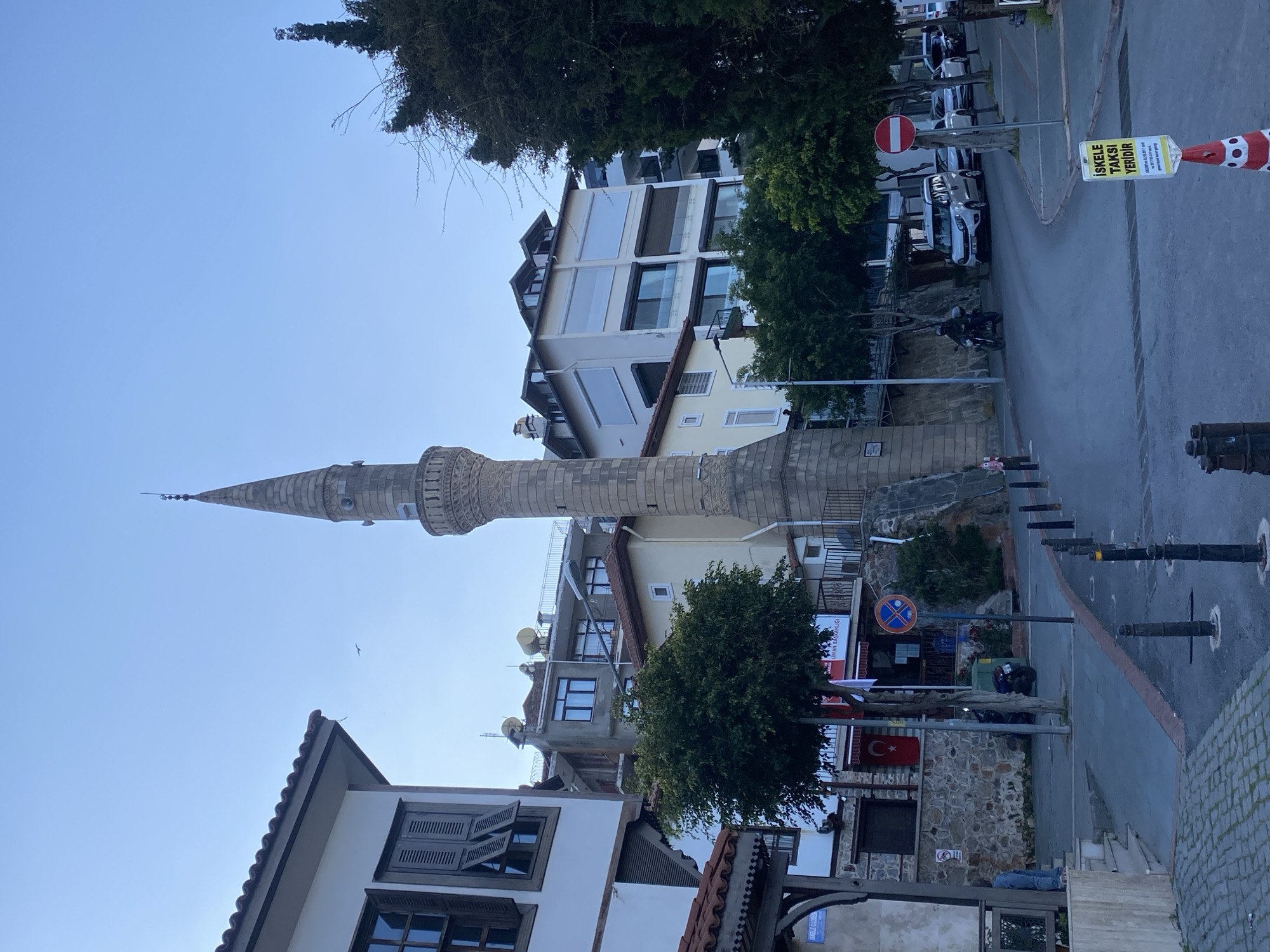 This media file presents a street scene in Alanya, Turkey, showcasing a blend of traditional and modern urban architecture under a clear, bright sky, likely in the late afternoon or early morning.
The central and most prominent feature is a tall, slender minaret, constructed of light-colored stone, featuring several ornate balconies (şerefes) and crowned with a conical spire. To the left of the minaret, part of a traditional-style building with a tiled roof and wooden shutters is visible. To the right and in the background, a mix of multi-story buildings with large windows and balconies line the street.
The paved street, with visible parked cars and a motorcycle, curves gently. Street furniture such as bollards and streetlights are present. A Turkish flag is prominently displayed on a building to the lower right. Several trees, including conifers and deciduous types, are scattered throughout the scene, adding greenery. Small birds can be seen flying in the distant sky.
Key location cues include the Turkish flag and a yellow sign with text in Turkish that reads "İSKELE TAKSİ YERİDİR," translating to "Pier Taxi Stand." A red circular traffic sign (likely 'no entry' or 'do not enter') is also visible. The scene is devoid of clear human activity, presenting a calm, static snapshot of an urban environment.