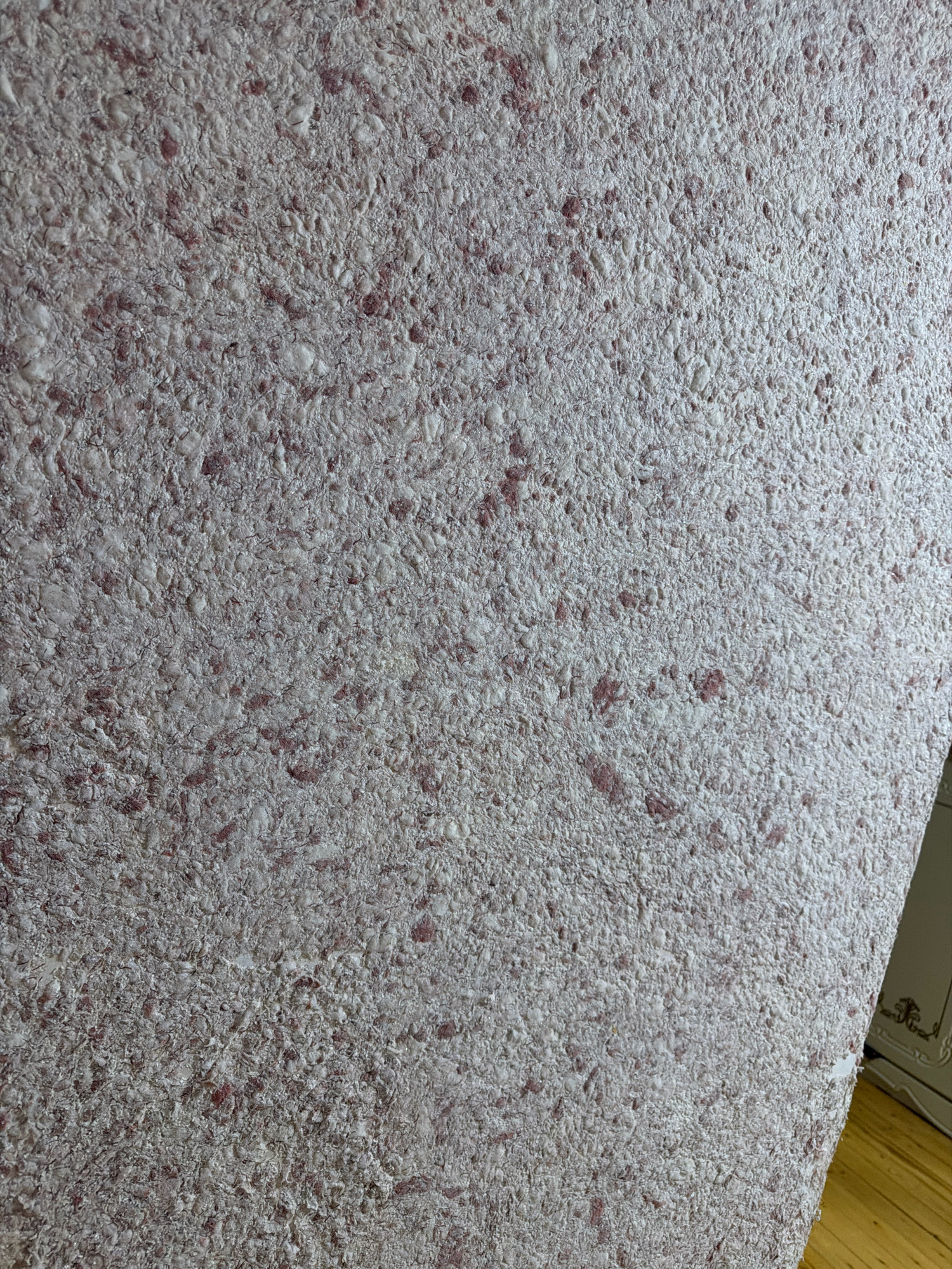 A close-up, high-angle shot shows a textured surface that appears to be wallpaper. The wallpaper is a light, neutral color, possibly off-white or pale gray, with small, irregular specks of pink or reddish-brown embedded within it. The texture is rough and uneven, with raised areas and depressions, giving it a tactile quality. The lighting is soft and diffused, casting no harsh shadows. The image does not contain any people, objects, actions, or interactions. The context of Inakri, Russia, is not visually represented in this image.