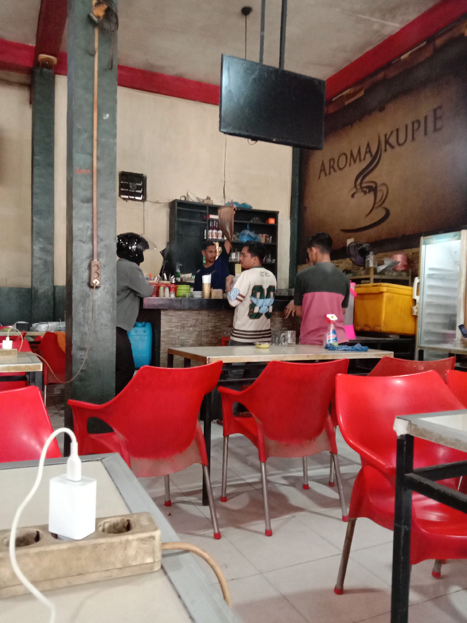 The image depicts an indoor scene of a modest coffee shop in Lhokseumawe, Indonesia. The primary focus is on the seating area with several bright red plastic chairs arranged around tables. In the background, there's a counter area where three individuals are present. Two men appear to be working behind the counter, one of whom is preparing something, while another man, wearing a helmet, stands near the entrance to the counter area. Another man is visible in the background, partially obscured. A large, vintage-style graphic of "AROMA KUPIE" with a steaming coffee cup logo adorns the wall. The environment suggests a casual, everyday setting for patrons to relax and enjoy beverages.