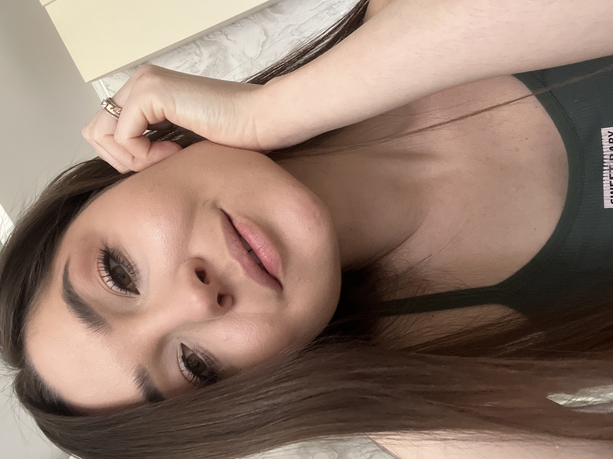 A woman with brown hair and brown eyes is looking at the camera. She is wearing a dark green tank top with the words "Sweet Baby" printed on the strap. Her left hand is touching her face, and she is wearing a ring on her ring finger. The background is blurry, but it appears to be a light-colored wall and a countertop. The location is Unknown, Unknown.
