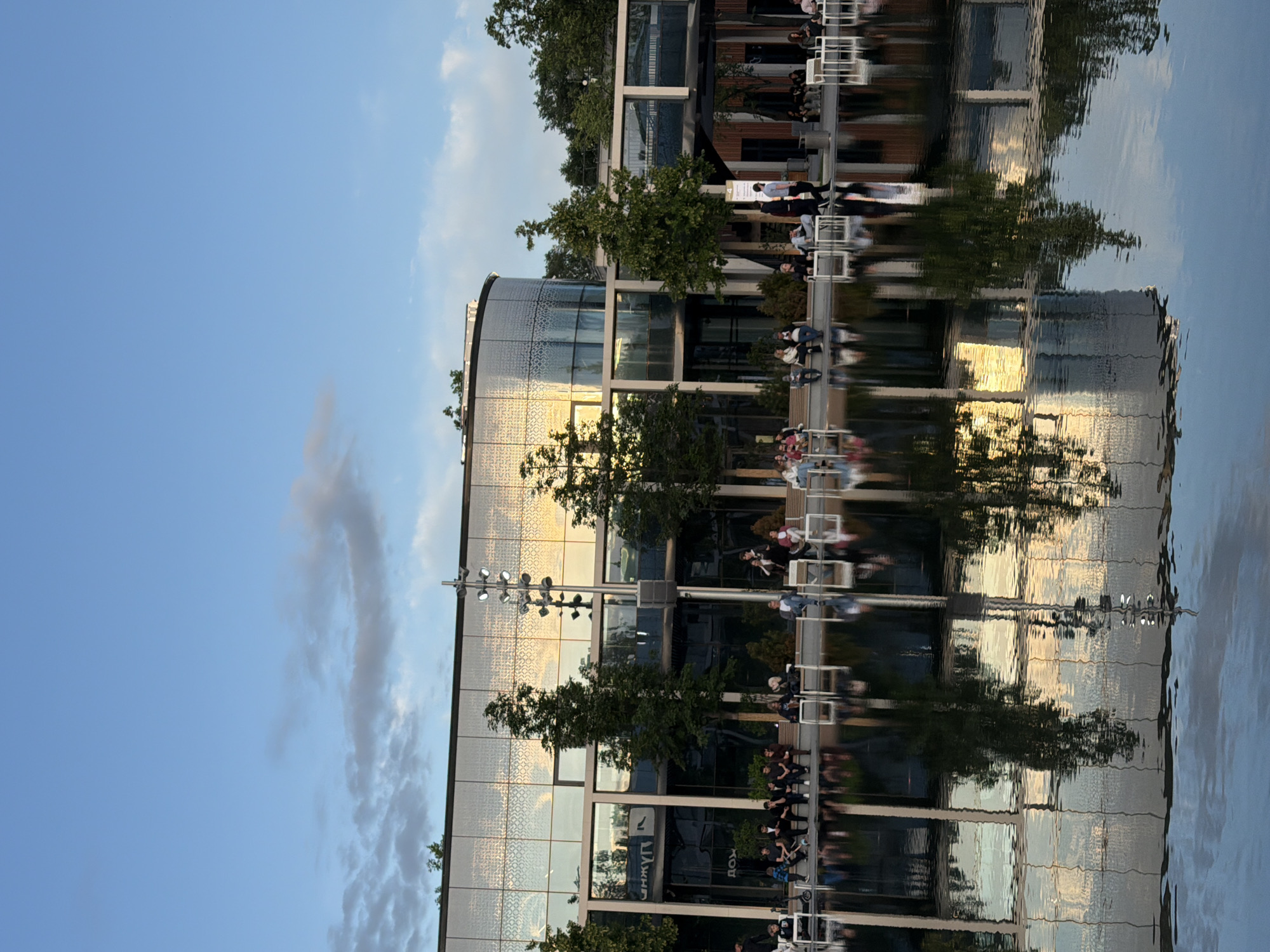 An outdoor scene depicts a modern building with a curved glass facade reflecting the sky and surroundings. In front of the building is a shallow body of water, possibly a reflecting pool, that mirrors the architecture and the sky. Several people are visible sitting on benches and chairs around the perimeter of the water, some facing the building, others engaged in conversation. Trees are interspersed between the building and the water. The sky is blue with scattered clouds, suggesting it is either early morning or late afternoon. The overall atmosphere is calm and relaxed, indicative of a public space or park.